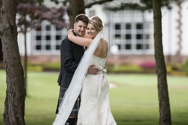 Cramond Kirk wedding: newlyweds embrace under trees, with the bride wearing a white gown and veil, and the groom in a dark suit, in front of a building with large windows.