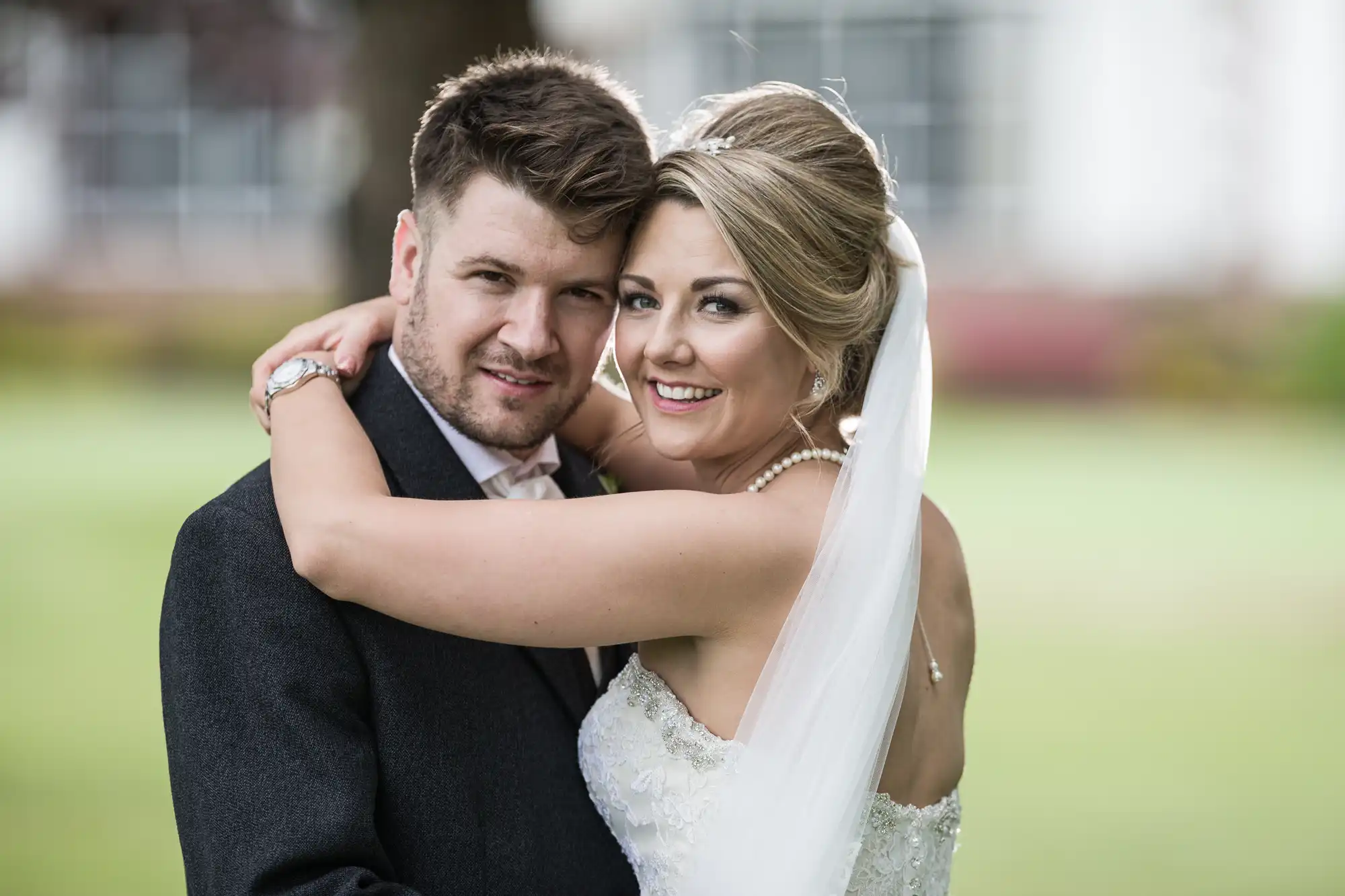 A bride in a white dress and veil embraces a groom in a dark suit, both smiling at the camera in an outdoor setting.