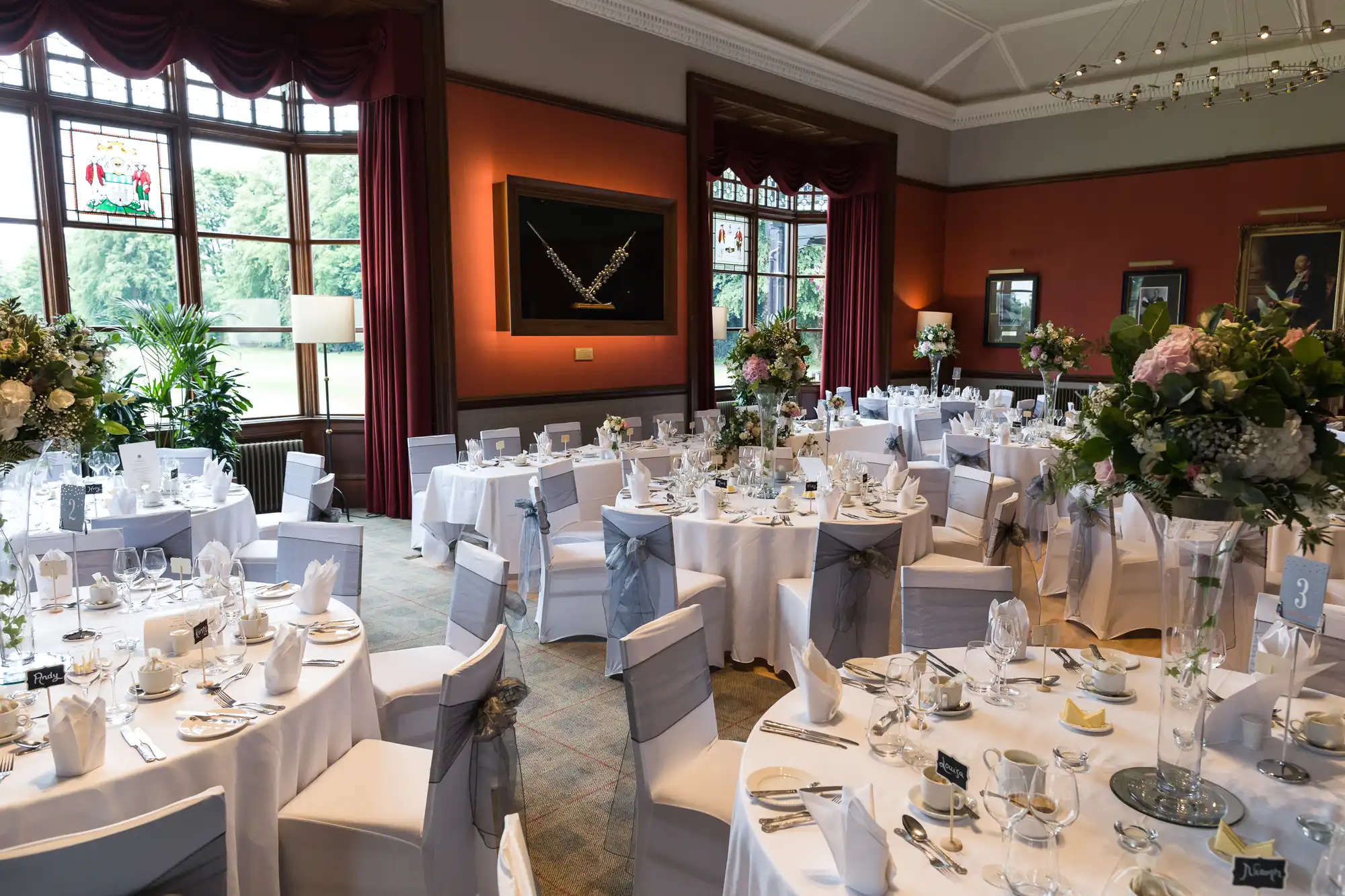 A formal dining room set up for an event with round tables covered in white tablecloths, floral centerpieces, and neatly arranged tableware. Large windows and framed artwork adorn the walls.