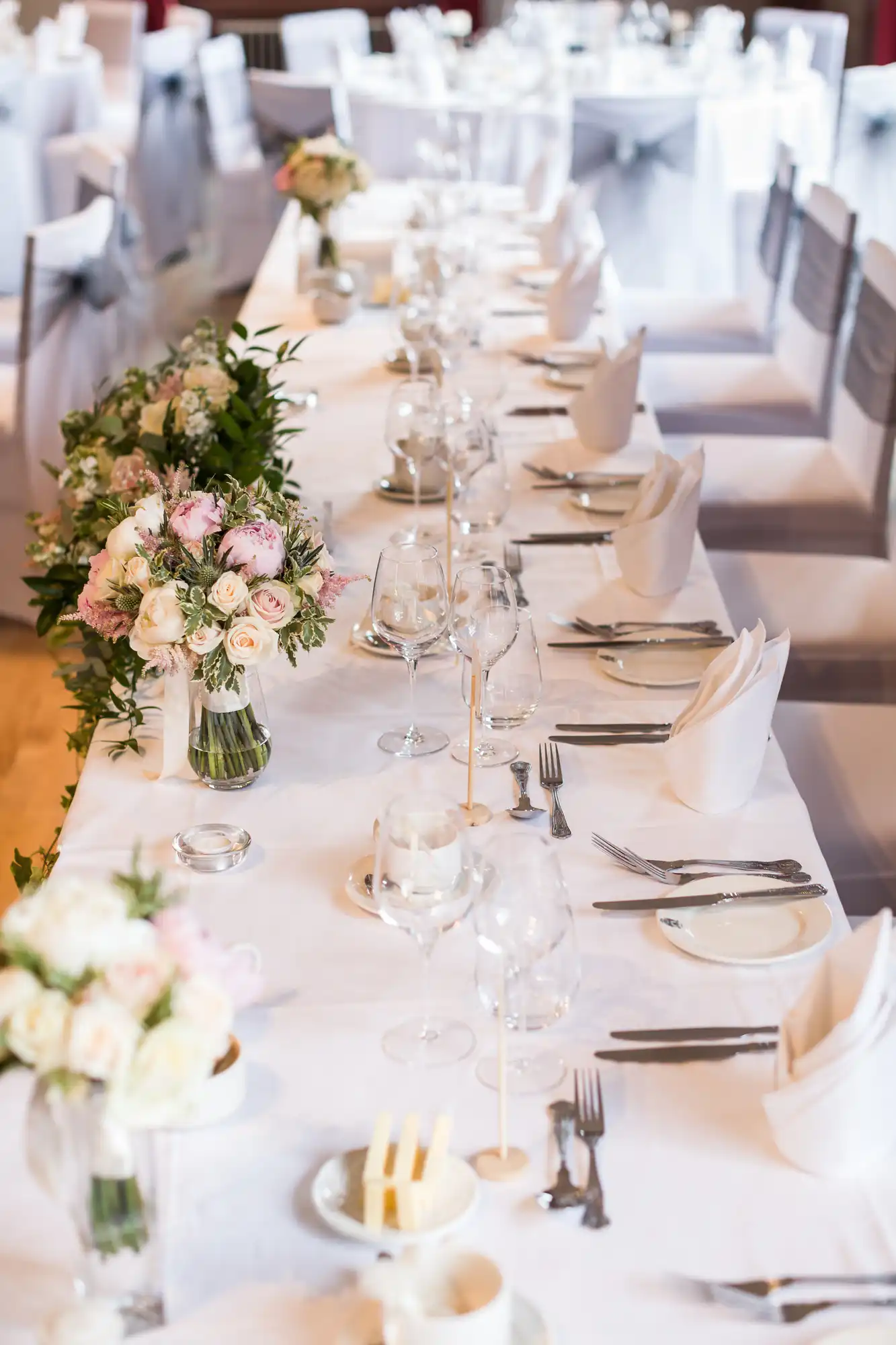 Elegant wedding reception table setup with white tablecloths, floral centerpieces, neatly arranged plates, glasses, and silverware, and white chairs adorned with grey sashes.