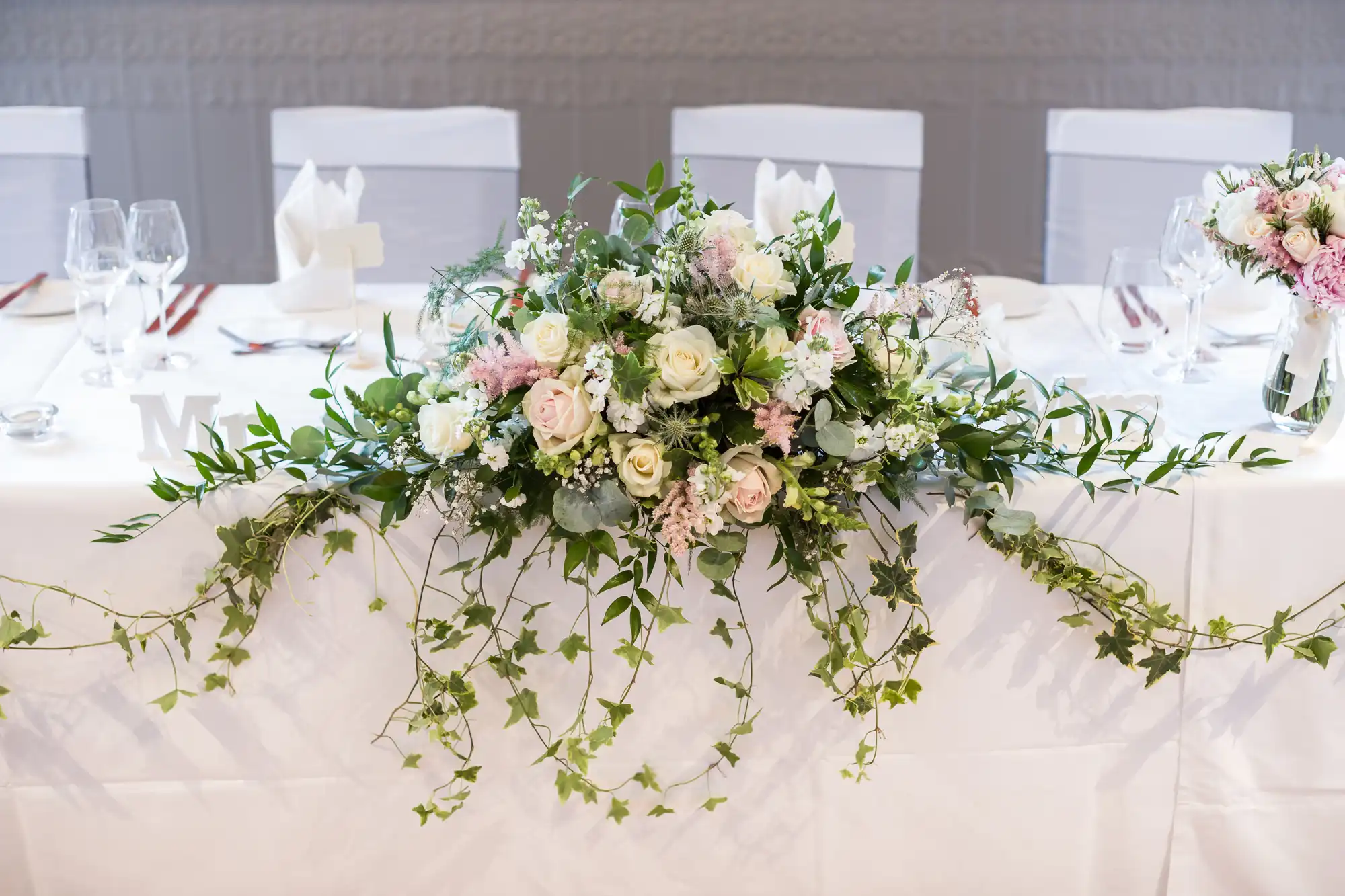 A white tablecloth-covered table is adorned with a floral centerpiece featuring pink and white roses, greenery, and trailing ivy. Crystal glasses and napkins folded in a standing position are also on the table.