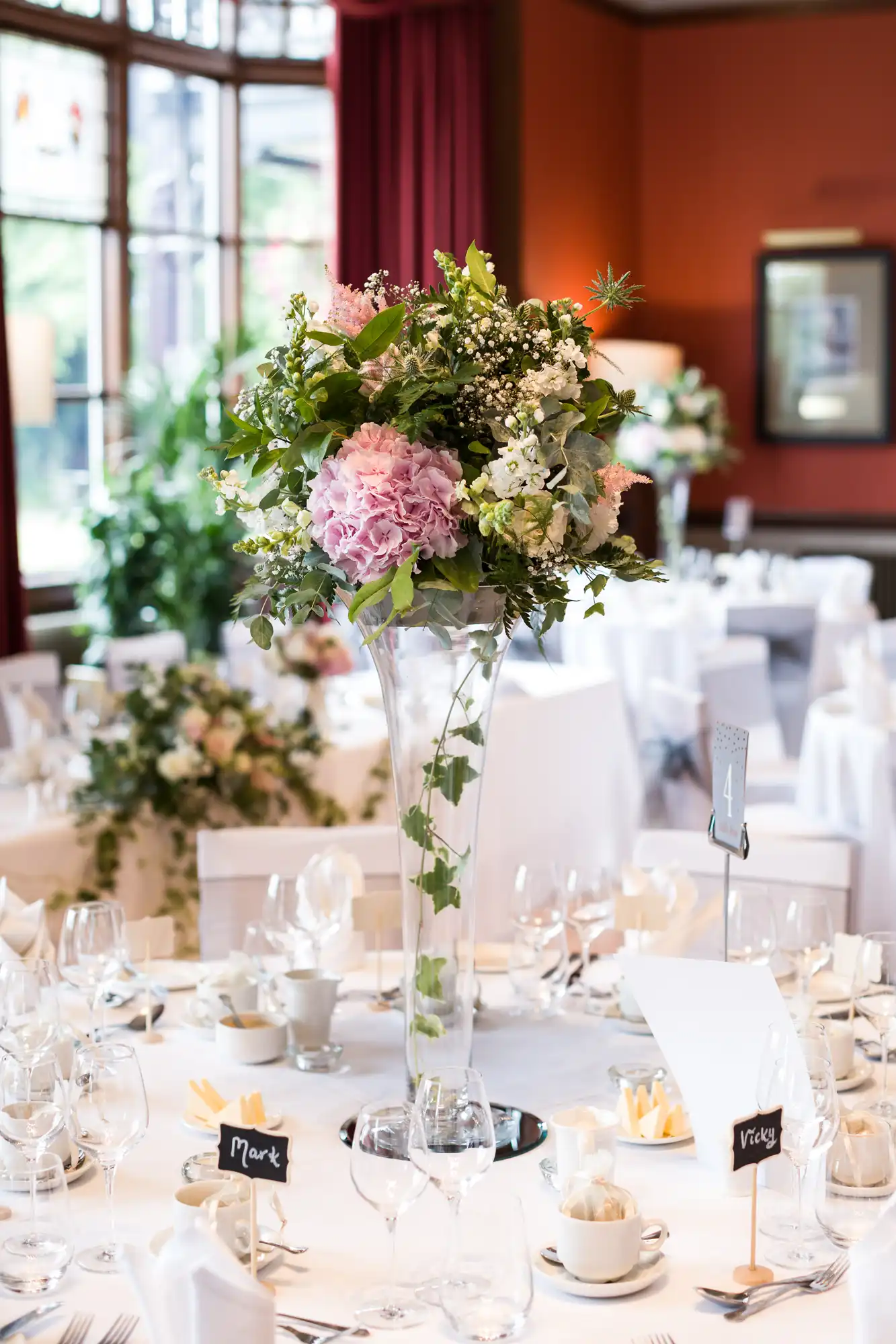 A wedding reception table with a tall centerpiece of pink and white flowers, surrounded by white table settings, name cards, and glassware.