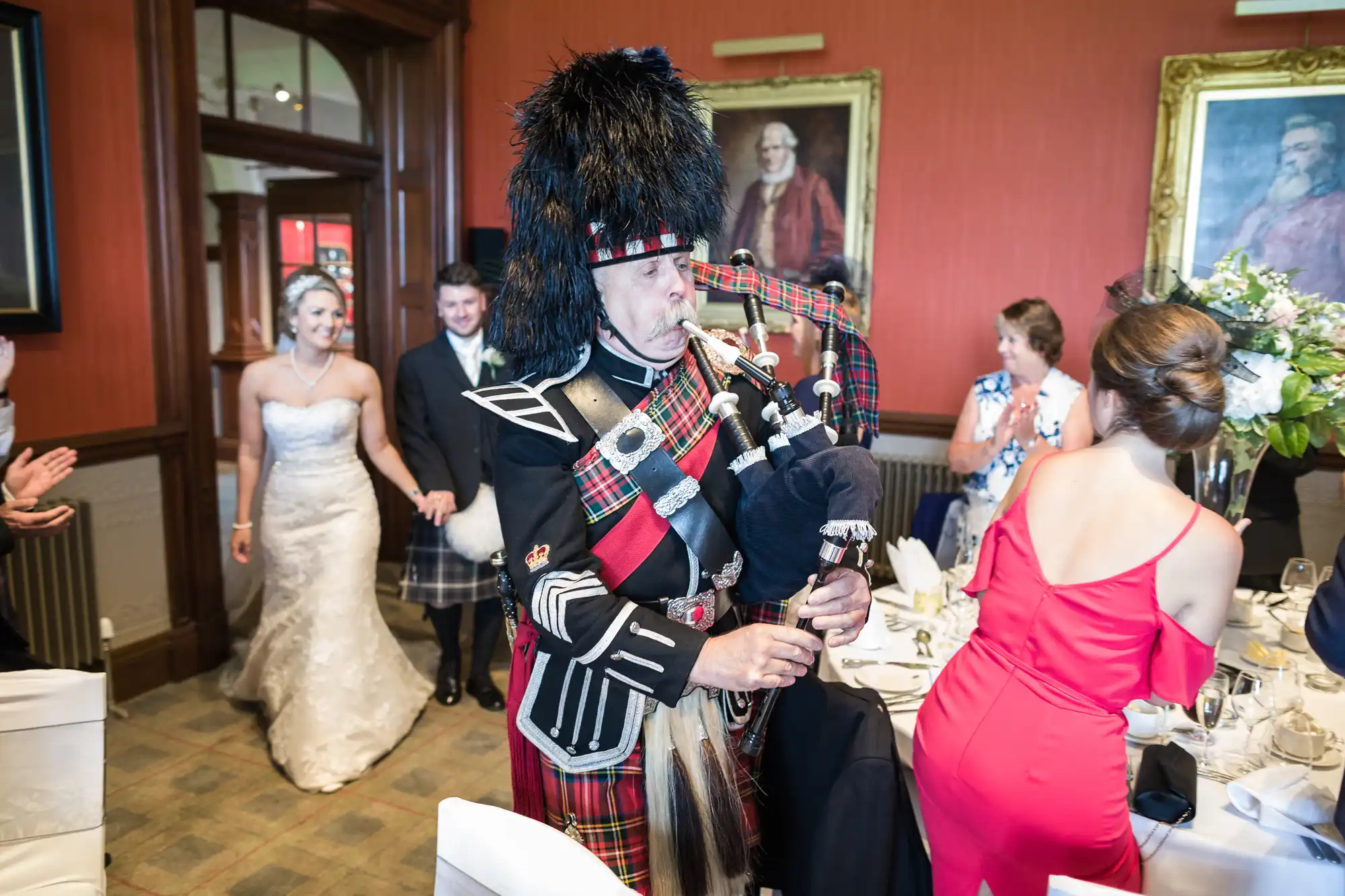 A bagpiper in traditional Scottish attire leads a bride and groom into a reception hall while guests applaud.