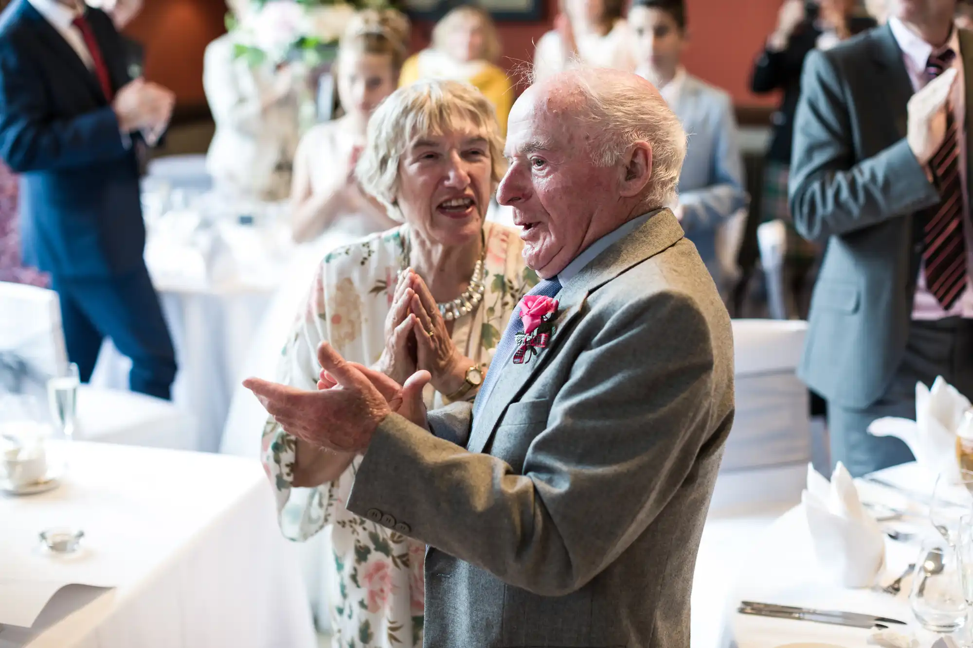 An elderly man and woman in formal attire are clapping and smiling in a brightly lit room with other people in the background.