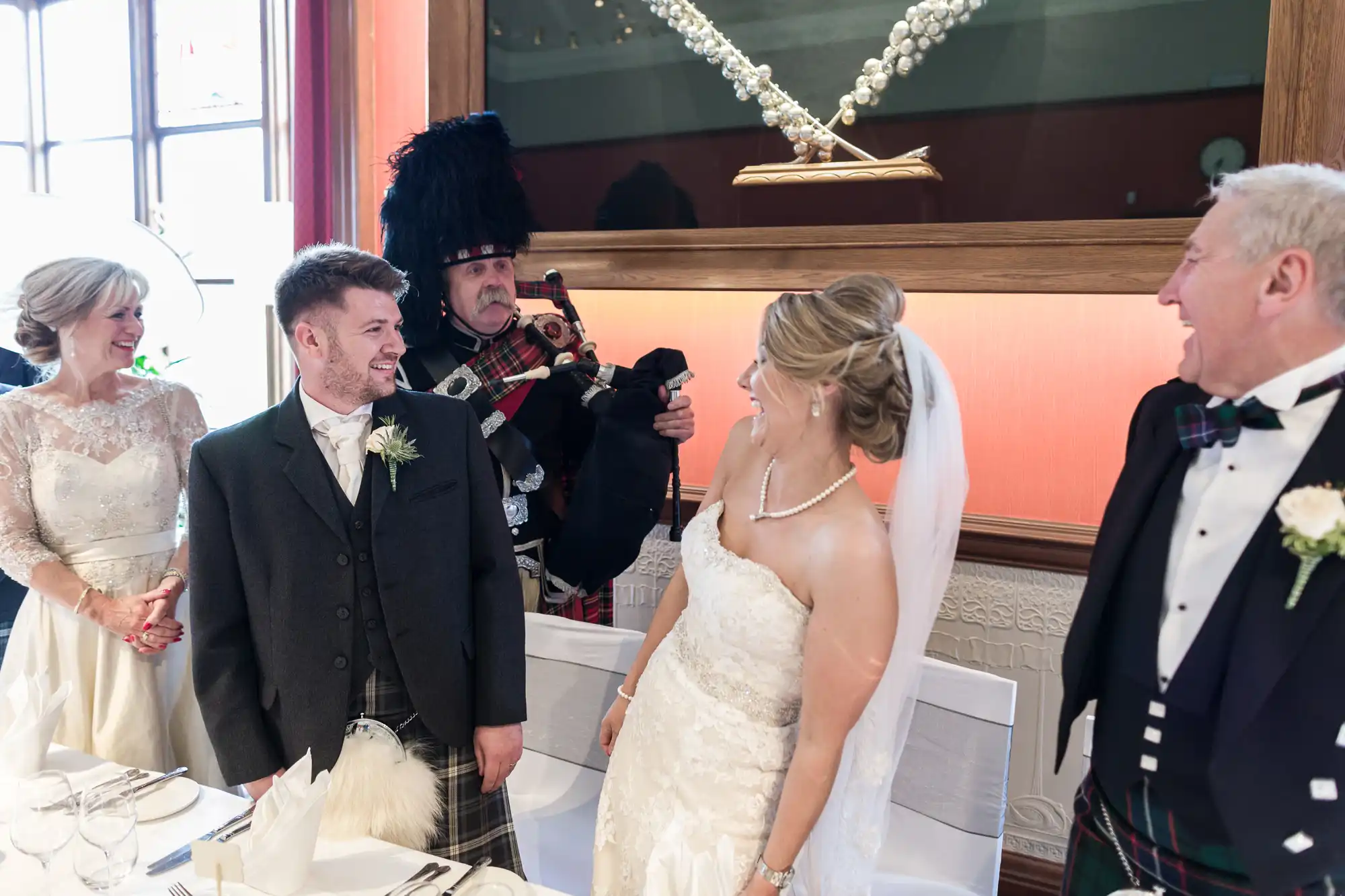 A bride and groom, dressed in wedding attire, laugh as a man in Scottish attire plays bagpipes. Three other individuals, also dressed formally, stand nearby, smiling and watching the bagpiper.