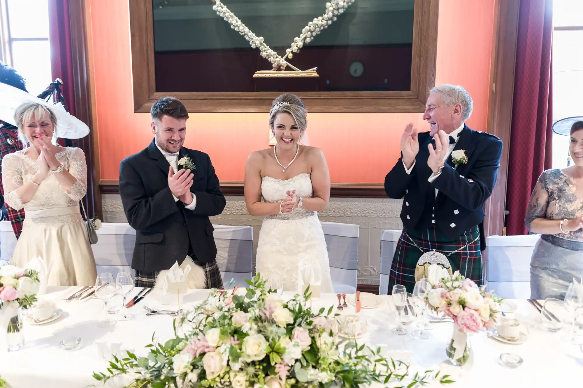 A bride and groom stand at a wedding reception table with their families. The bride and groom are clapping, and guests around them also appear to be clapping and smiling.