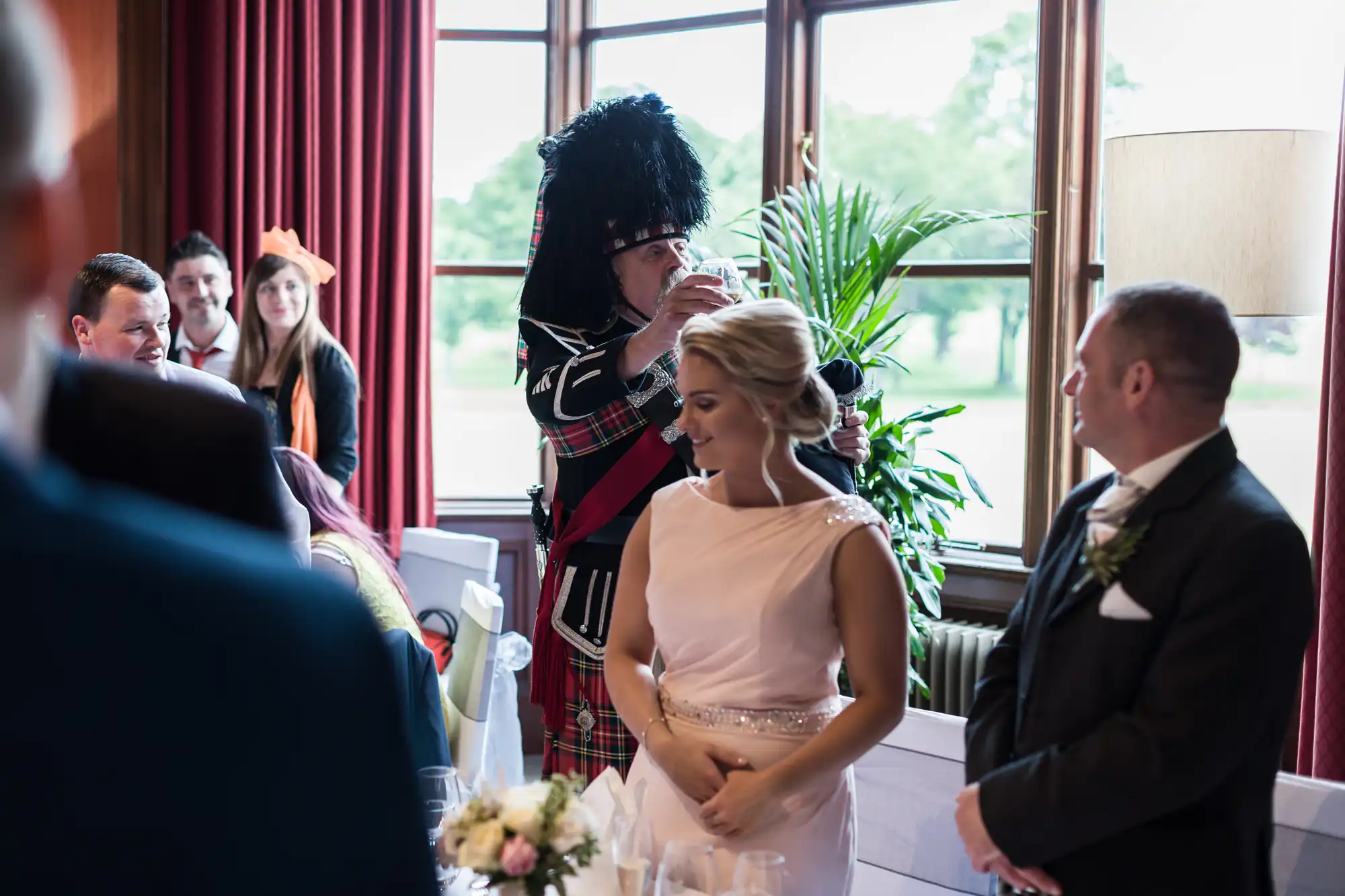 A person in a kilt and a feathered hat plays a bagpipe at an indoor gathering, with other attendees standing, one in a formal suit and another in a light pink dress, near a window with a green outdoor view.