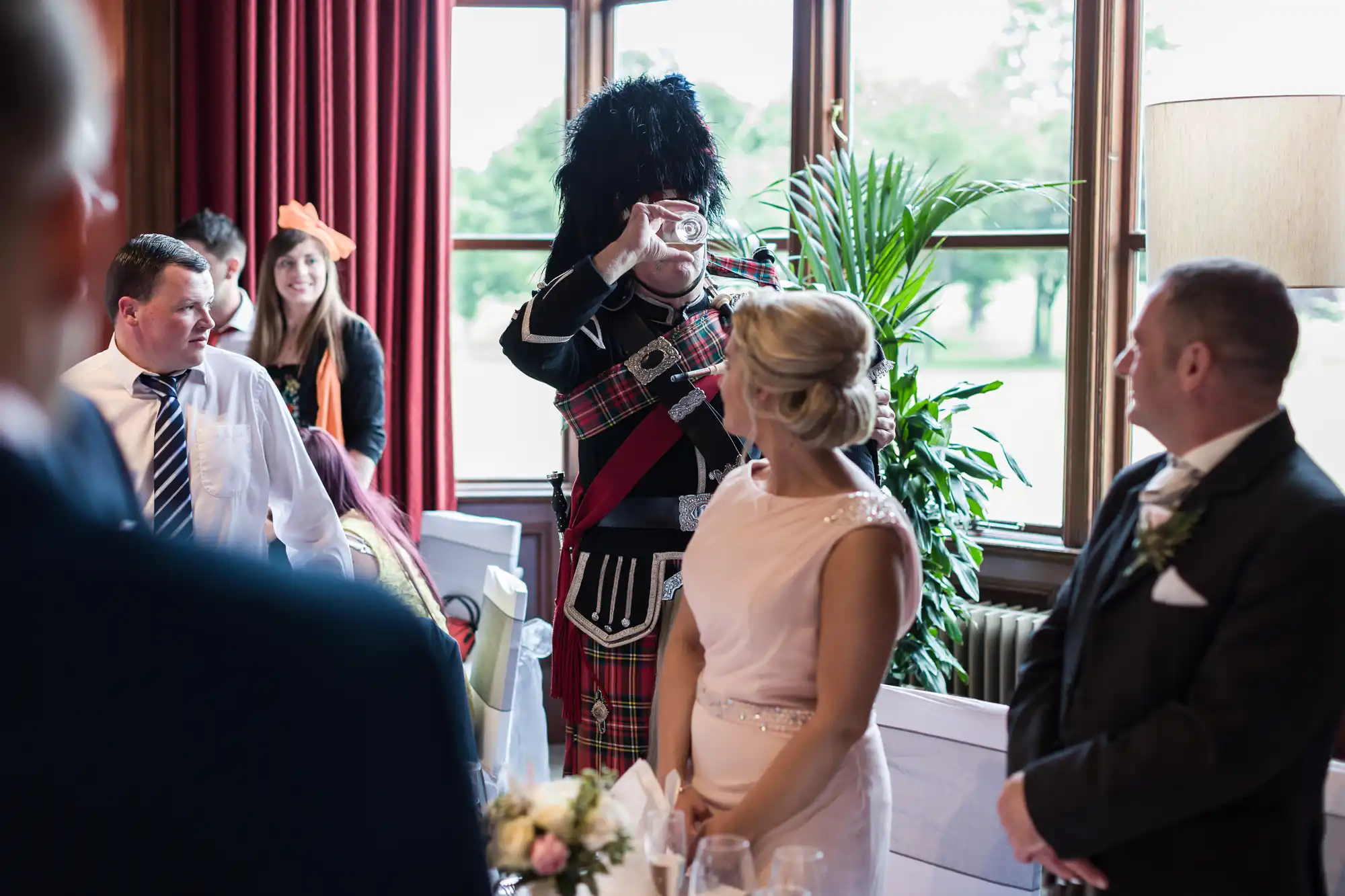 A man in Scottish traditional attire drinks from a glass while standing among a group of formally dressed people in a room with red curtains and large windows.