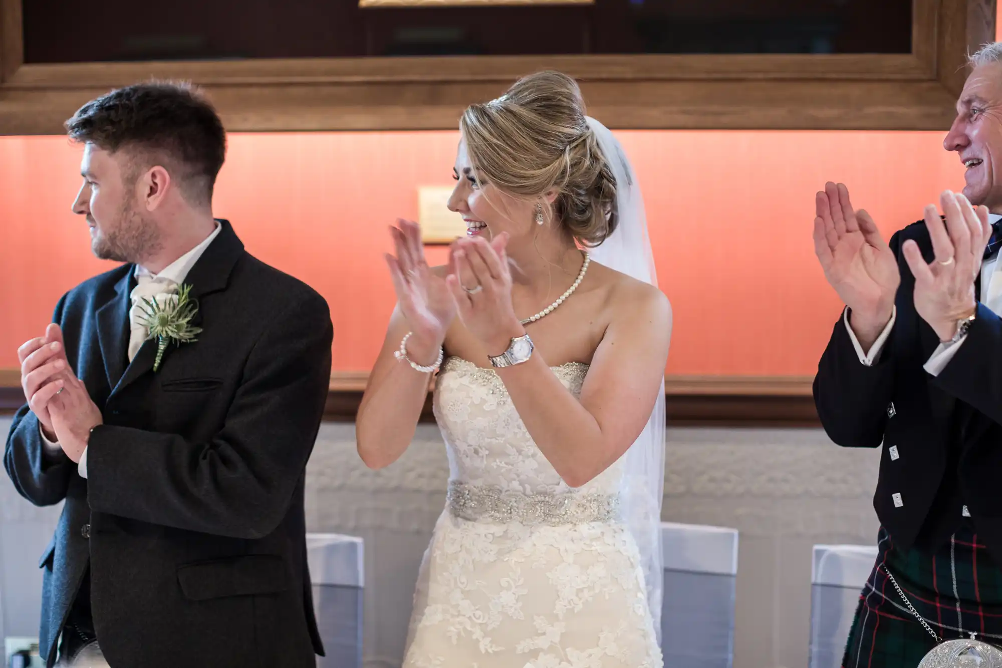 A bride in a lace wedding dress and veil, flanked by two men in formal attire, claps her hands while looking to her left.