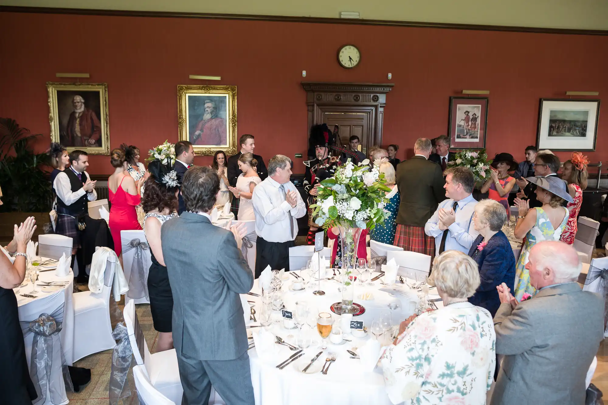 A group of people dressed formally are standing and clapping around dining tables adorned with white tablecloths and flower arrangements in an ornate room. Paintings and a clock are visible on the walls.