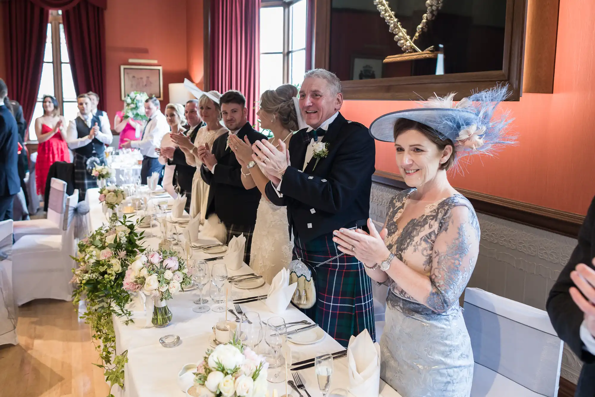 Guests in formal attire stand and clap around a long, elegantly decorated table at a wedding reception. Flowers and folded napkins adorn the table. .