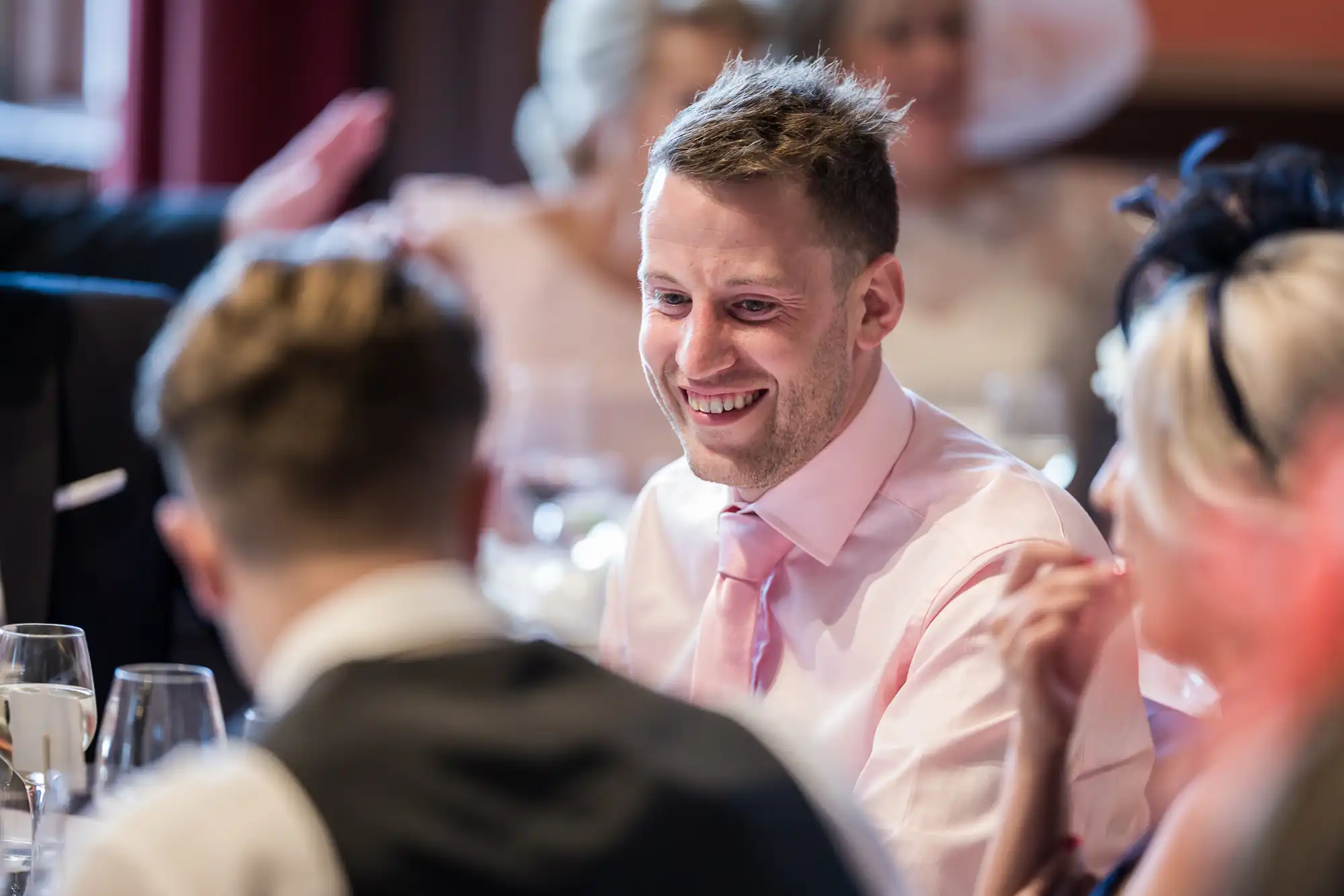 A man in a pink shirt and tie smiles while seated at a formal event, engaging in conversation with people around him.