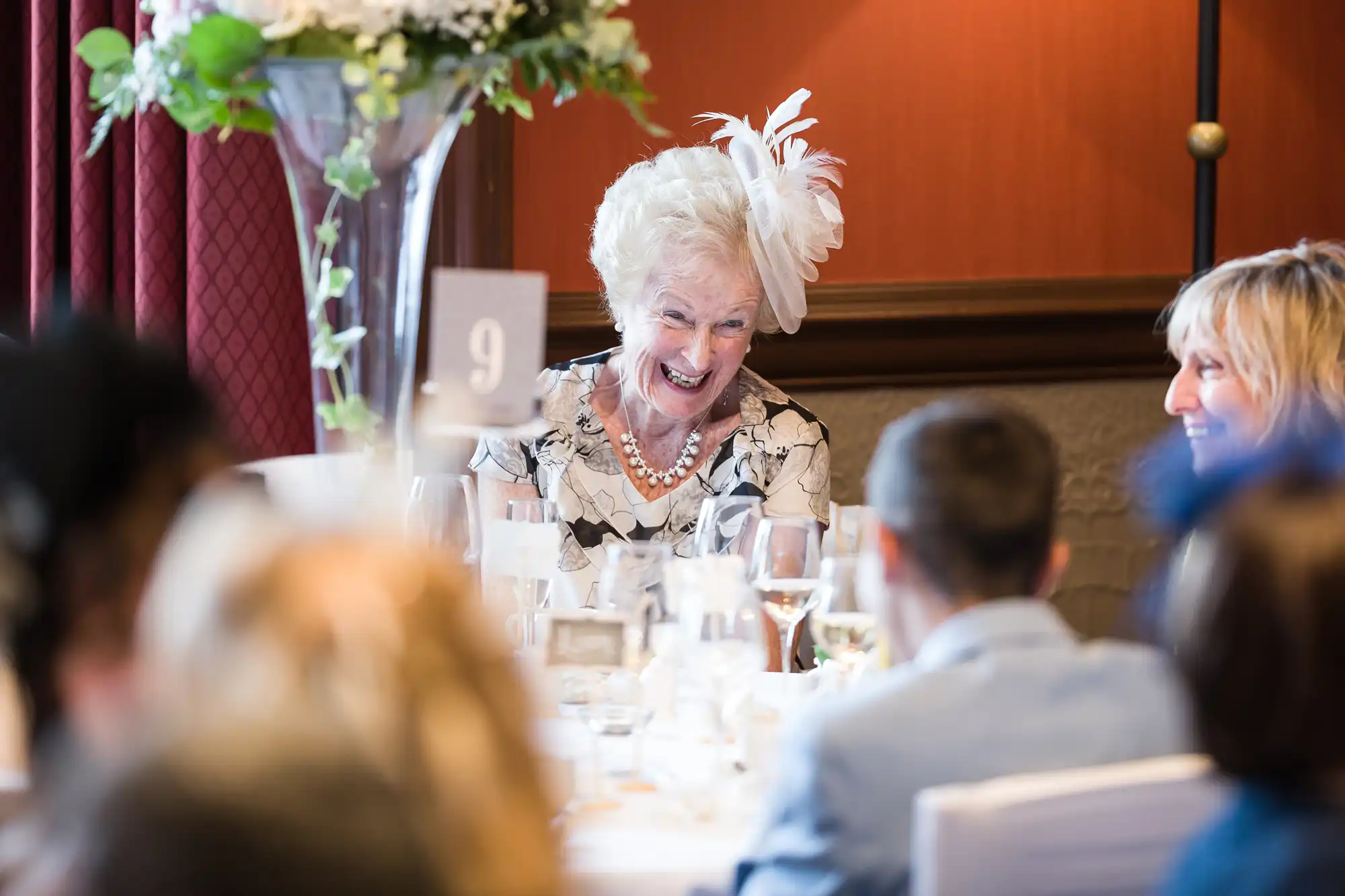 Elderly woman with white hair and fascinator smiling and laughing at a dining table set for an event, with several people seated around her.