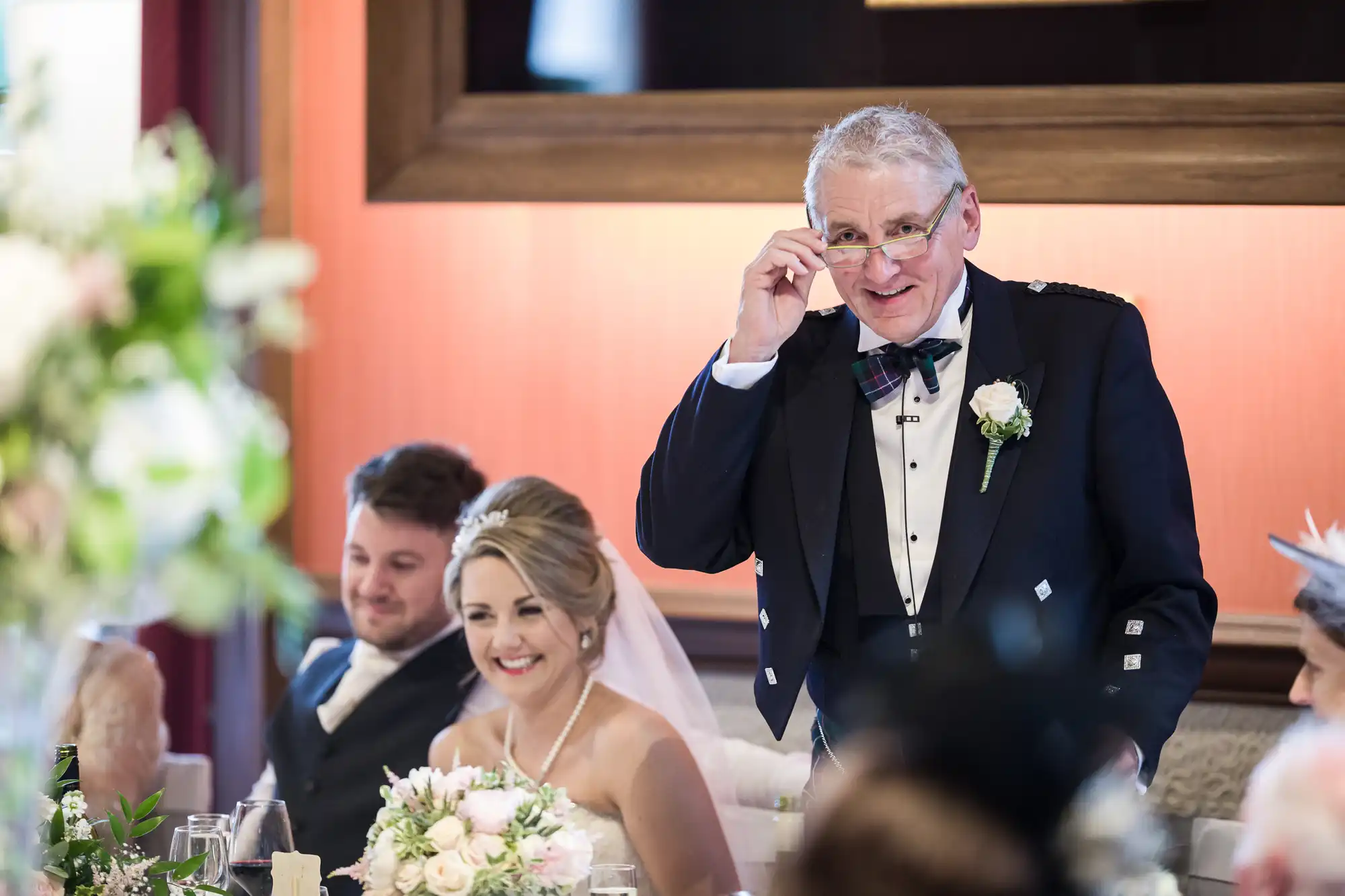 An older man in formal attire speaks at a wedding reception, adjusting his glasses. A bride in a white gown and groom sit smiling at the table decorated with flowers. Guests are visible in the background.