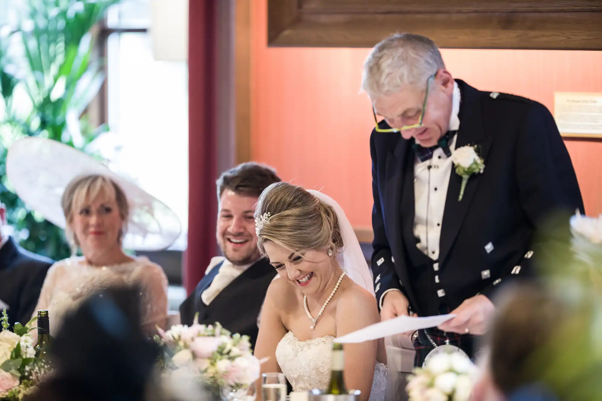 A bride and groom sit at a table smiling during a wedding reception while a man standing beside them reads from a piece of paper. A woman in a hat is seated nearby.