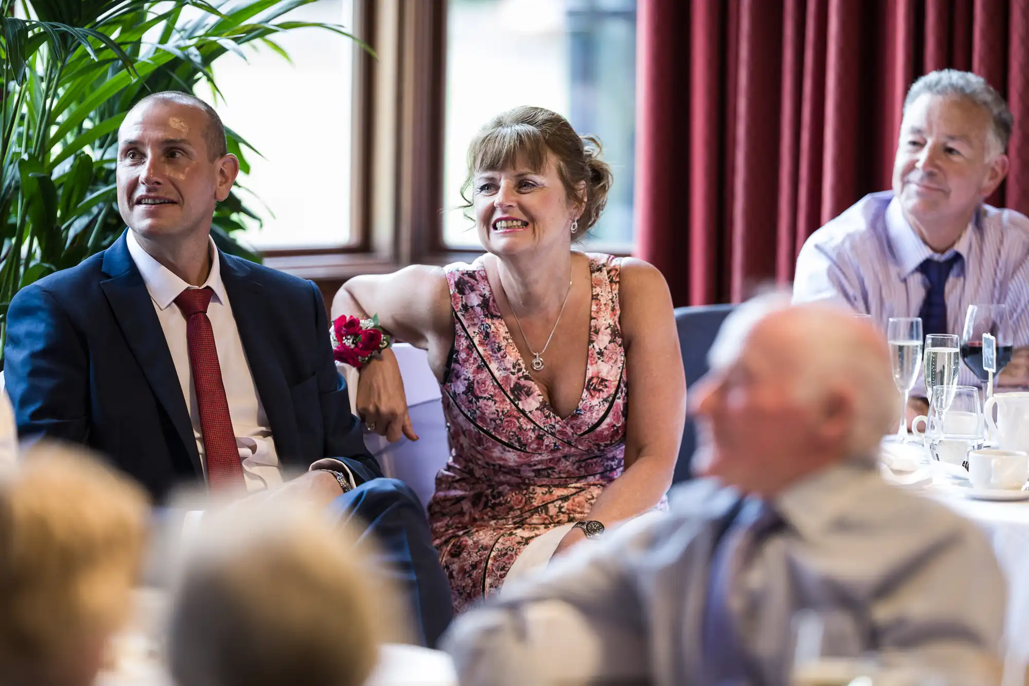 A group of people, including a man in a suit and a woman in a floral dress, sit at a table, smiling and looking towards the same direction, during an indoor event.