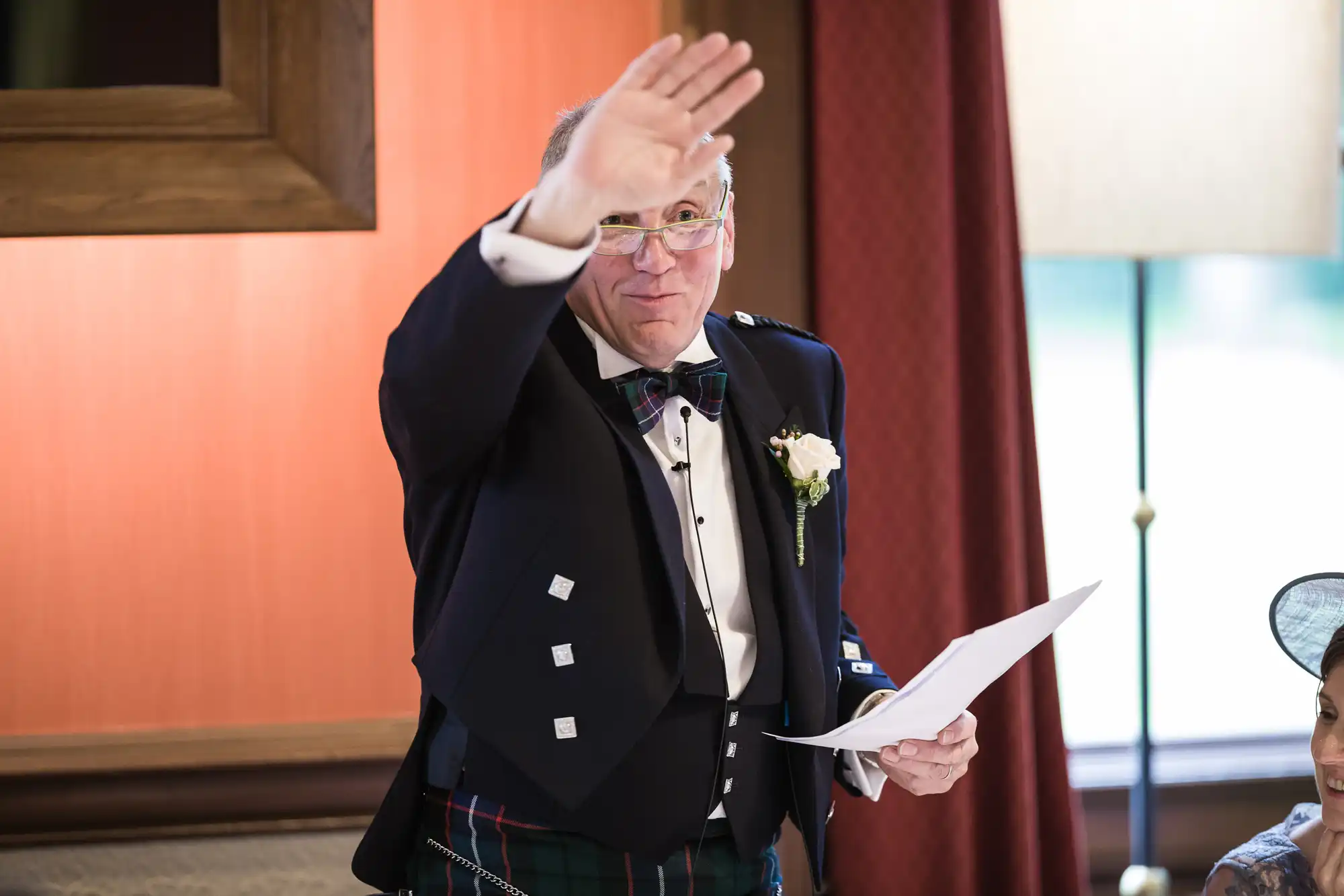 A man wearing a formal kilt outfit is speaking and gesturing with one hand raised while holding a piece of paper in the other. He stands in an indoor setting with red walls and a lamp in the background.