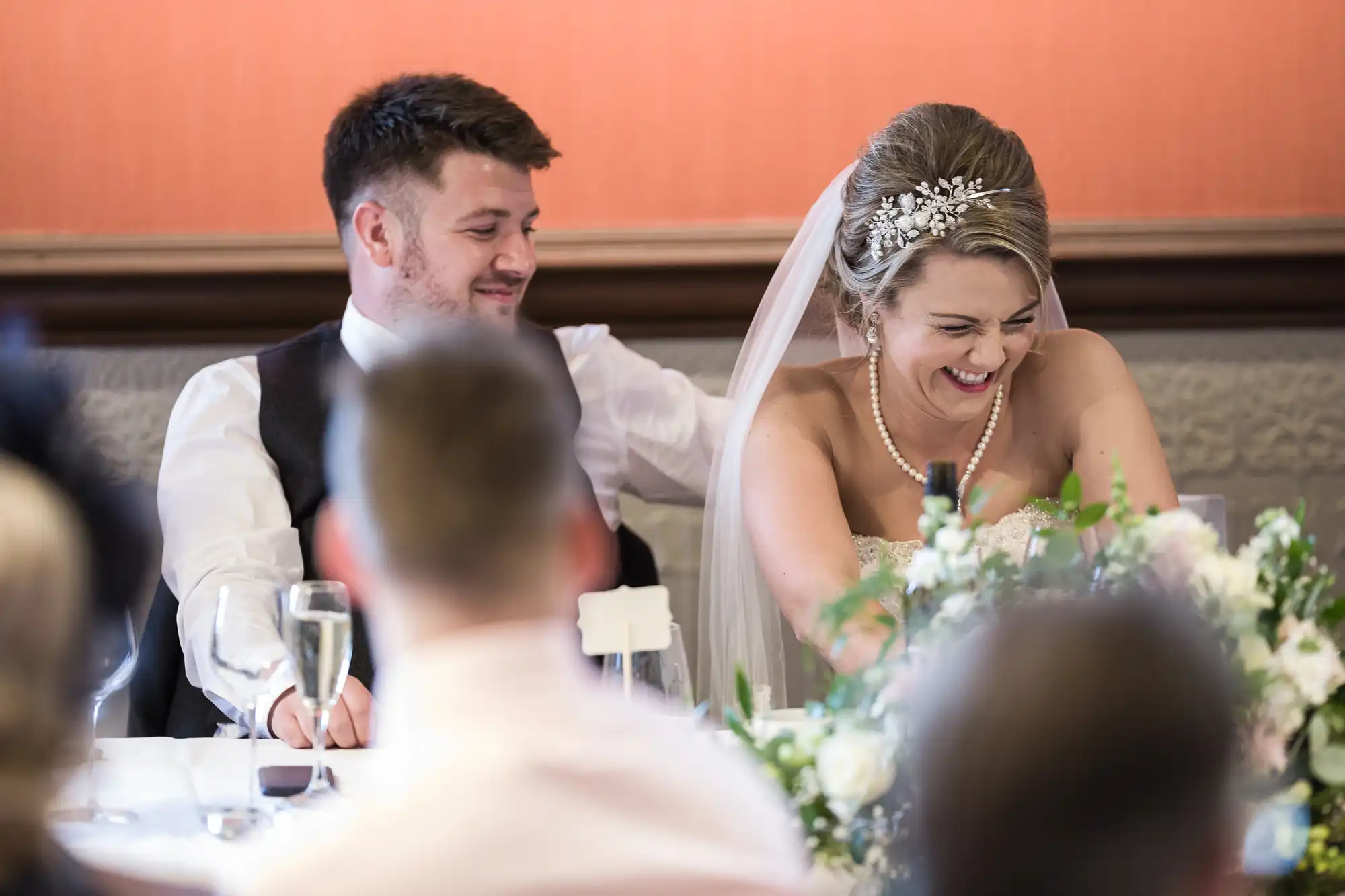 A bride and groom sit at a table during their wedding reception. The bride is laughing while the groom smiles at her.