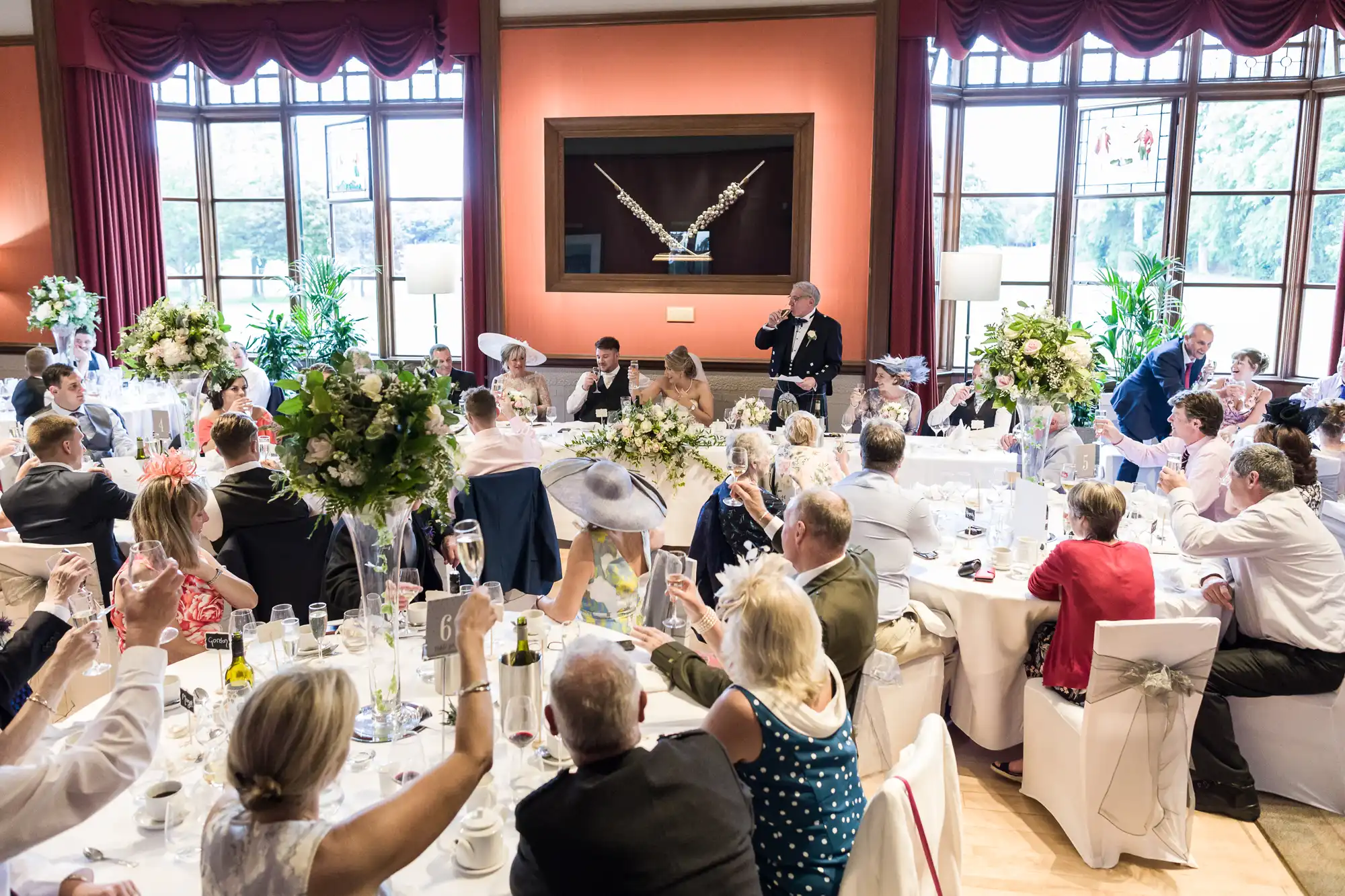 A formal gathering in a large, well-lit room with round tables set for a meal, featuring a speaker addressing attendees who appear to be toasting. Large windows and floral centerpieces are present.