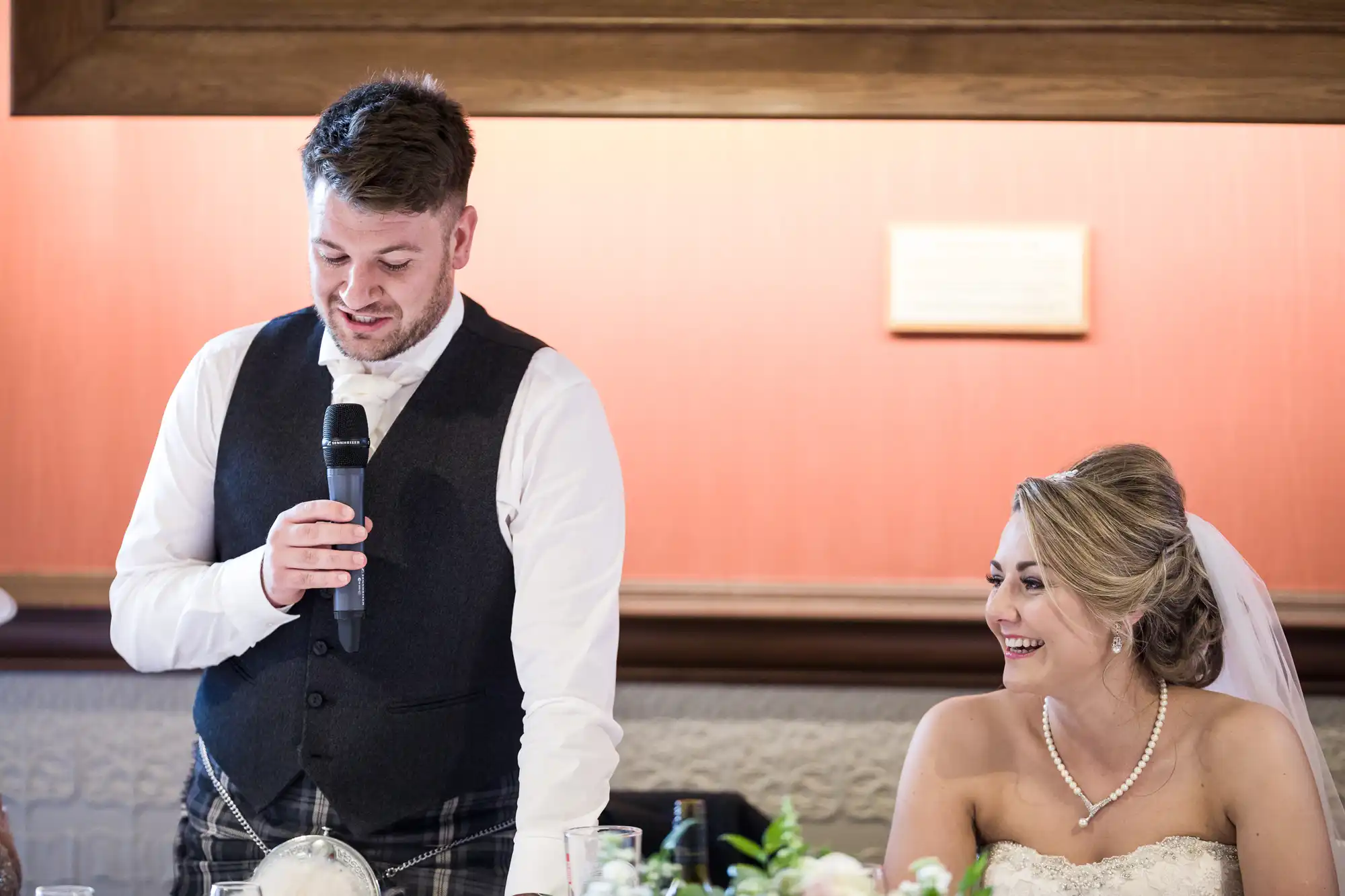 A man in a suit and tie is speaking into a microphone, while a woman in a wedding dress and veil sits beside him, smiling. They are indoors with a wooden frame and orange wall in the background.