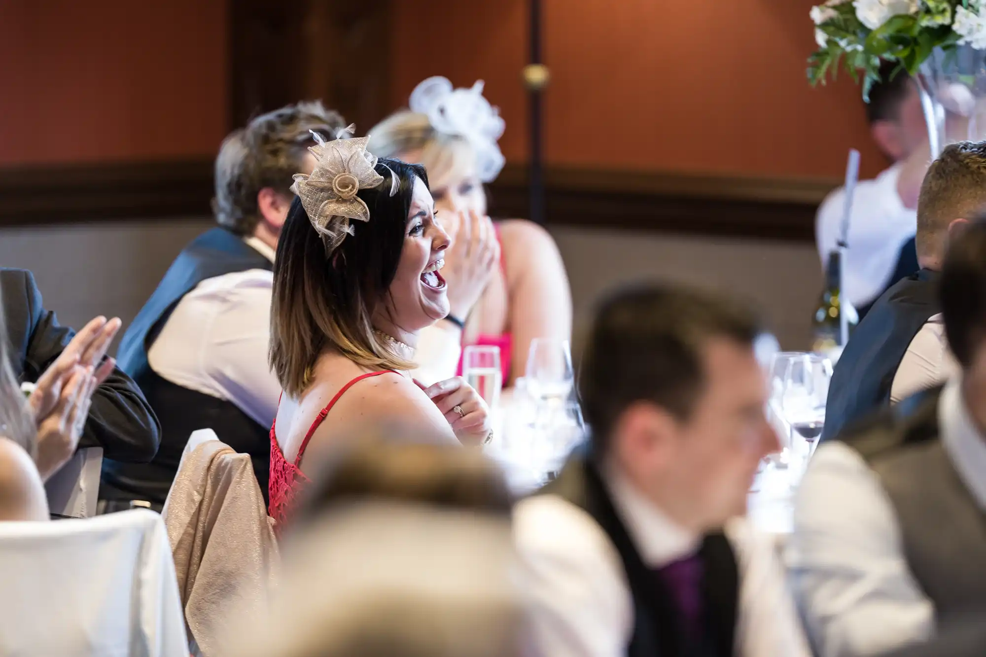 A woman wearing a fascinator sits at a table, smiling and looking to her right, at an event with other attendees seated and clapping in the background.