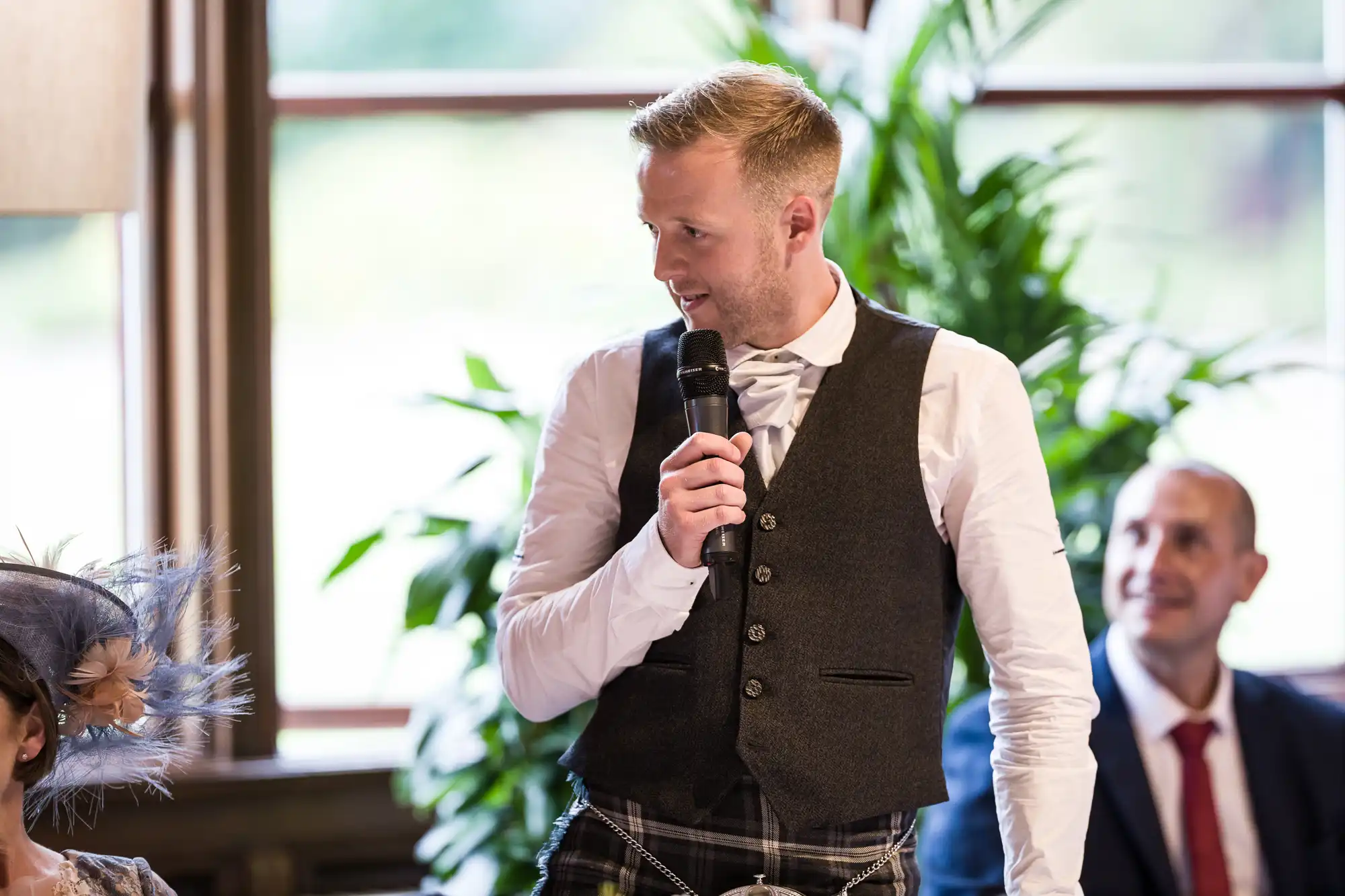 A man in a vest and tie speaks into a microphone at an indoor event, with greenery and large windows seen in the background.