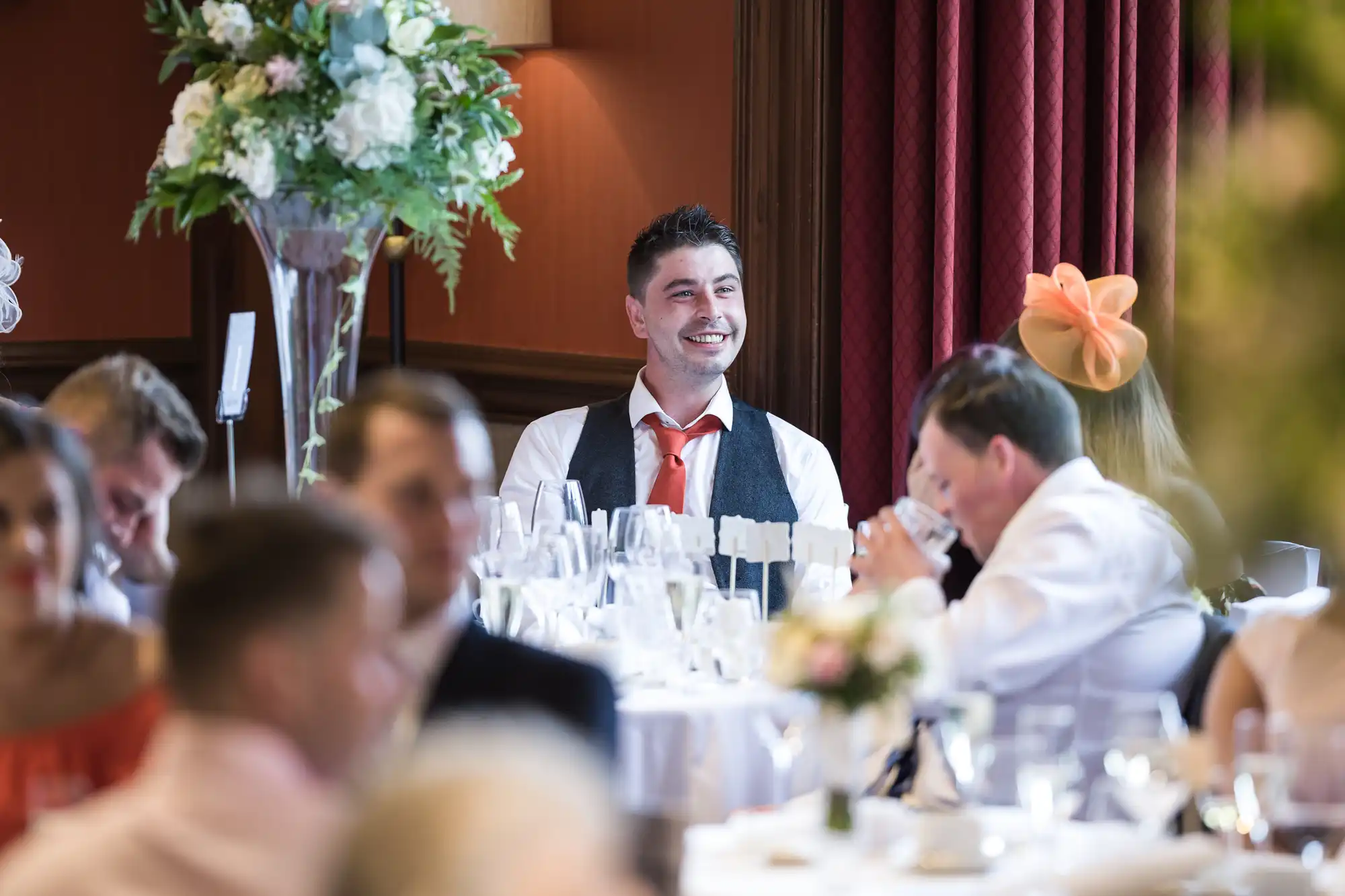 A man in a white shirt and tie sits at a table decorated with flowers, smiling at a formal event as people around him engage in conversation.