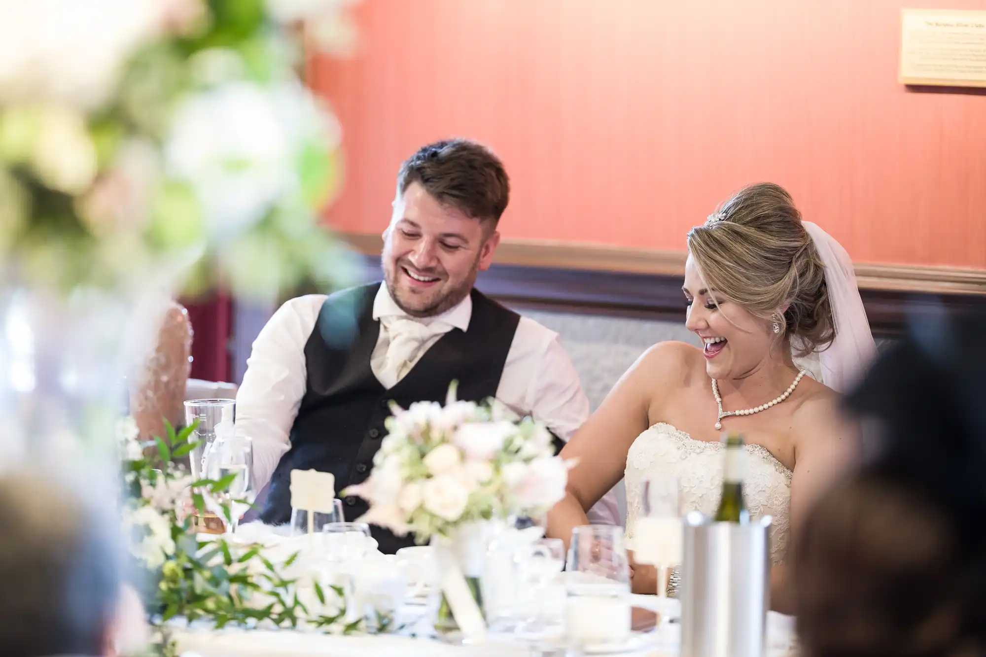 A groom in a black vest and a bride in a white dress and veil share a laugh while seated at a table decorated with flowers and tableware.