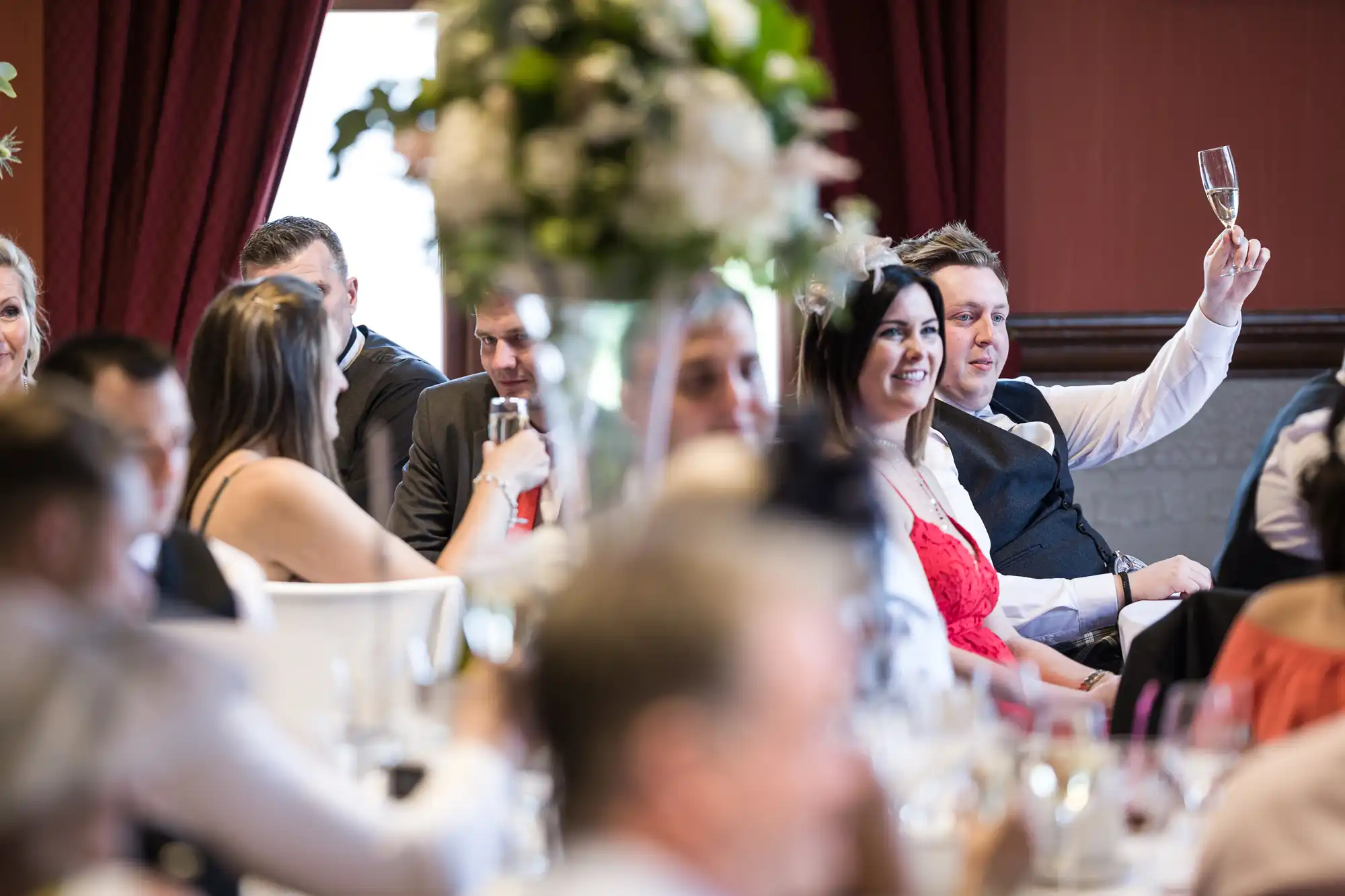 People seated at tables in a restaurant or event hall, with a man raising a glass in a toast, and others engaged in conversation.