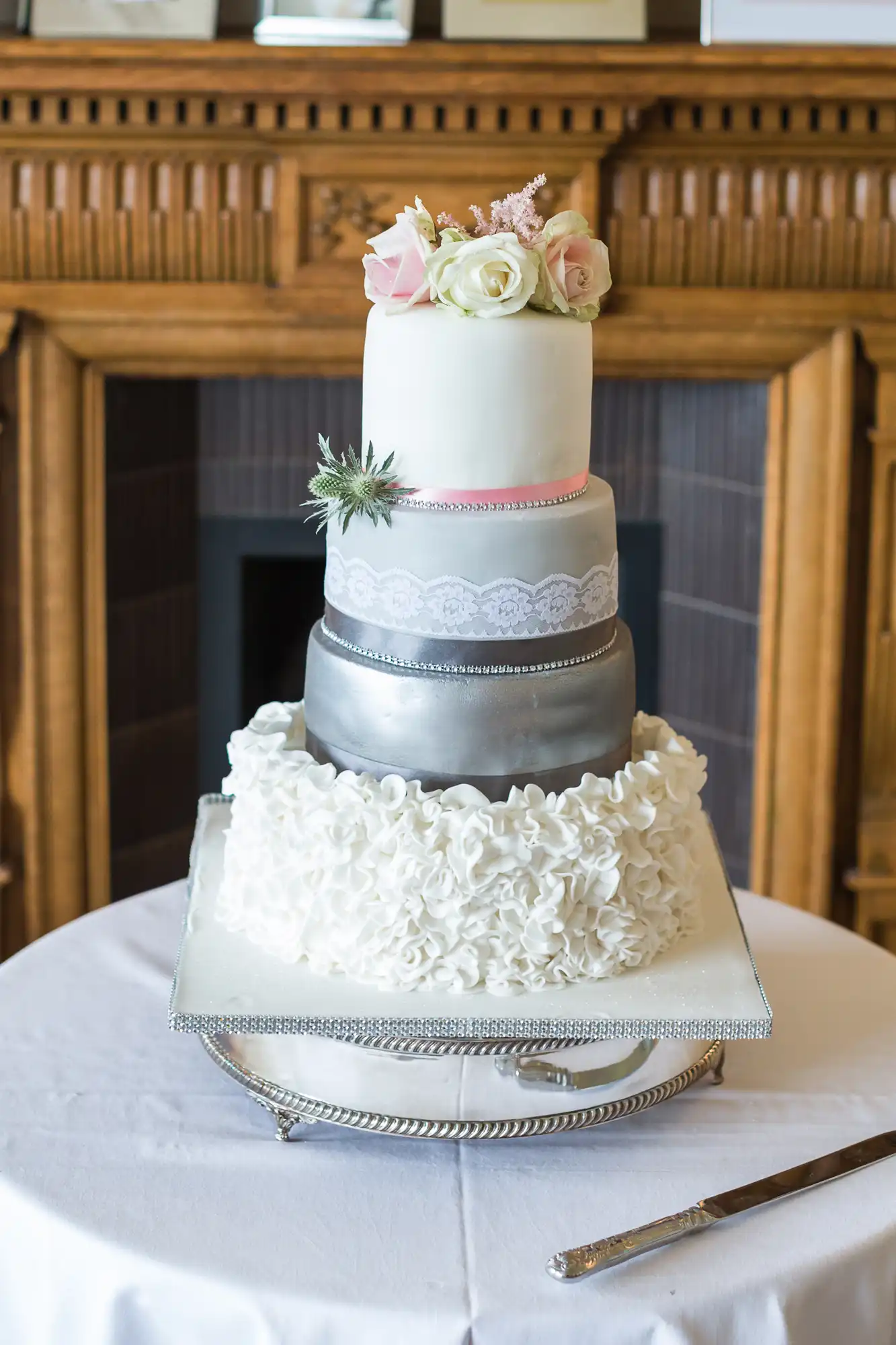 Three-tiered wedding cake with white, silver, and pink layers, adorned with lace, flower decorations, and placed on a cake stand on a white tablecloth beside a cake knife.