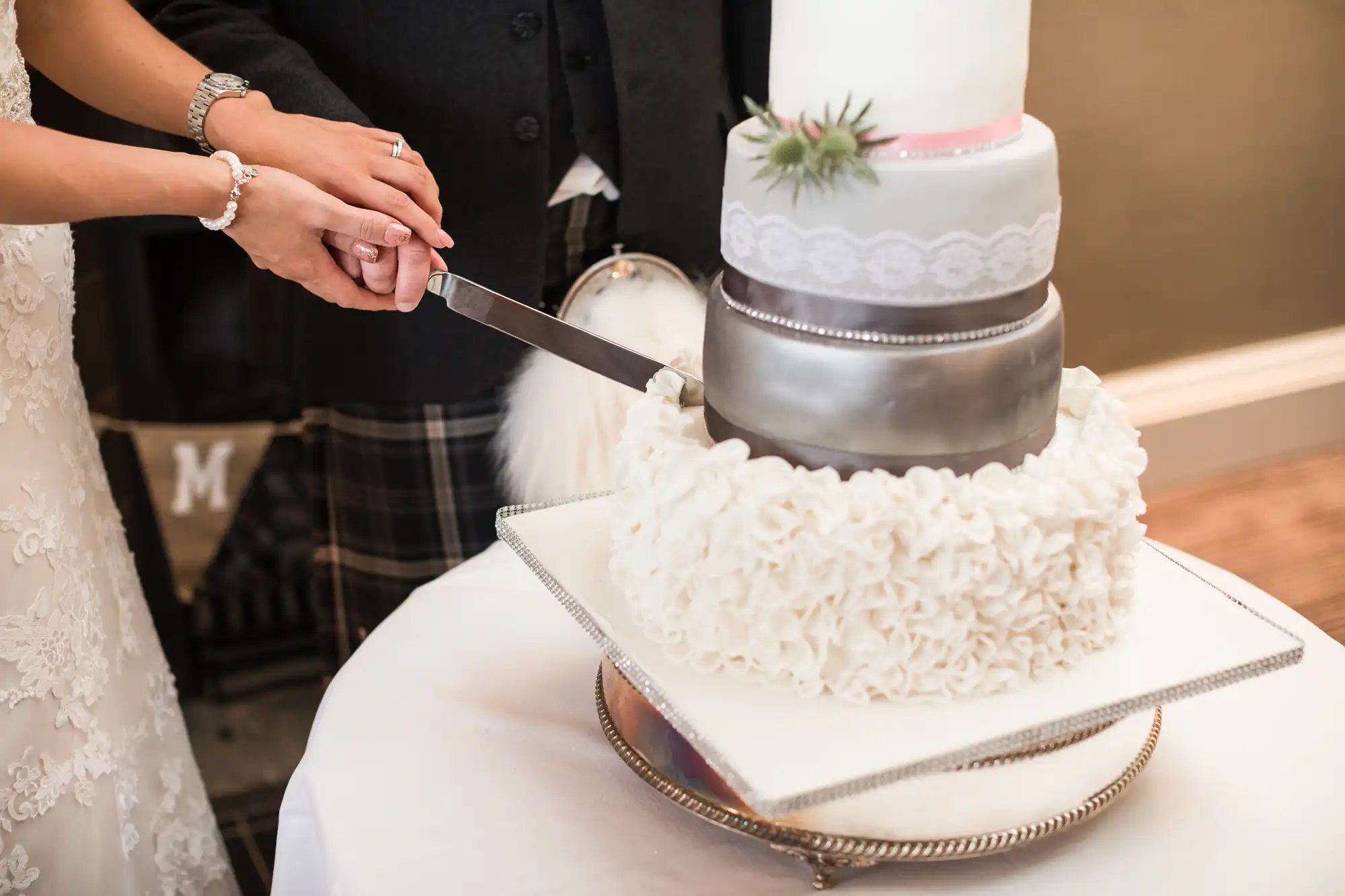 A couple cuts a white, multi-tiered cake adorned with lace and a small succulent on a table.