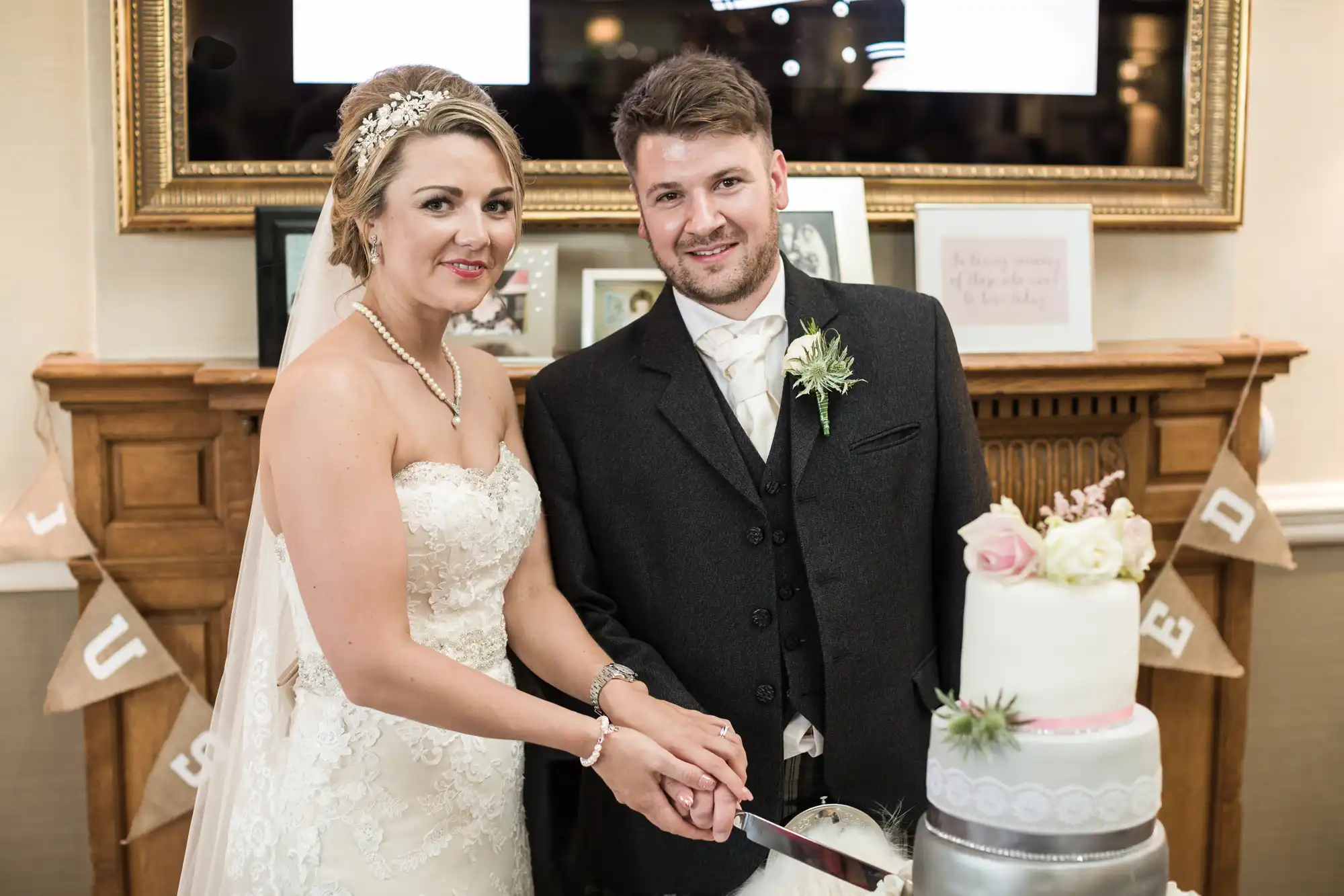 A bride and groom, both dressed in wedding attire, pose while cutting a three-tiered white wedding cake. The bride holds the knife, and a "Just Married" banner is visible in the background.