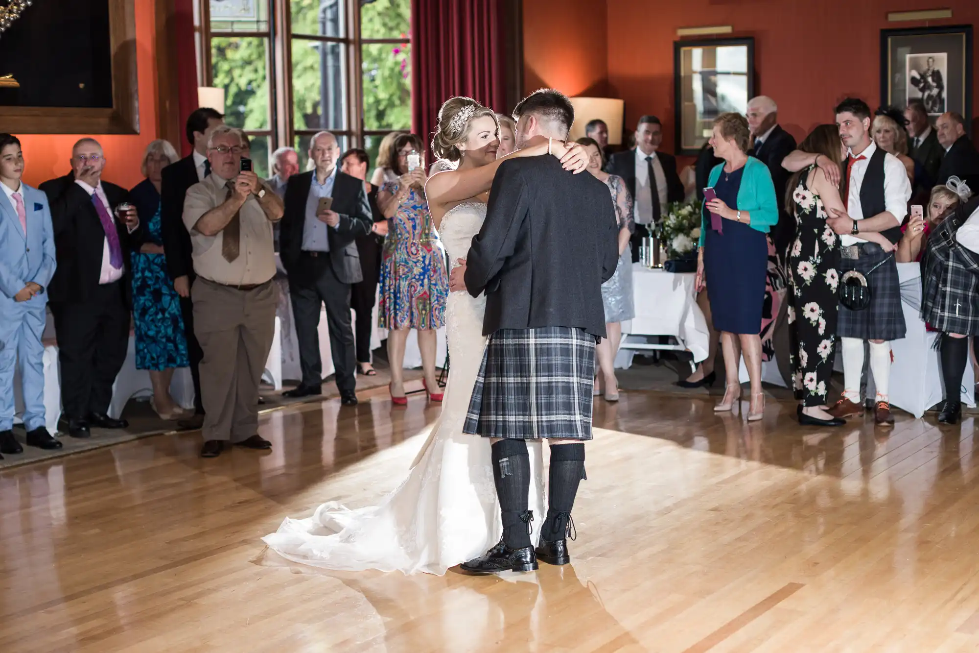 A couple, dressed in wedding attire, dances while guests watch at a wedding reception in a brightly lit hall.