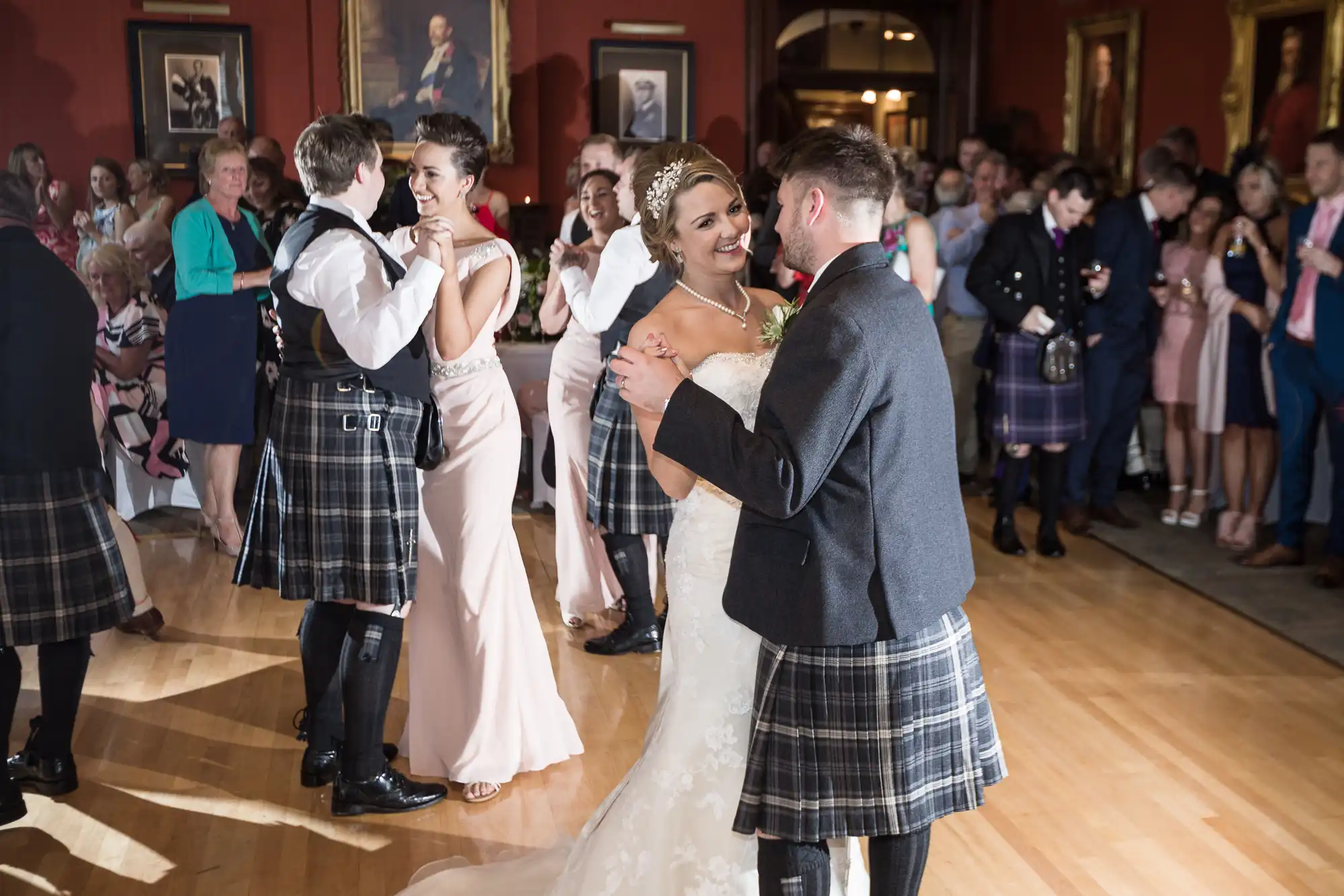 A bride and groom dance at their wedding reception, surrounded by guests, some clothed in traditional Scottish attire. Other couples are also dancing in the background.