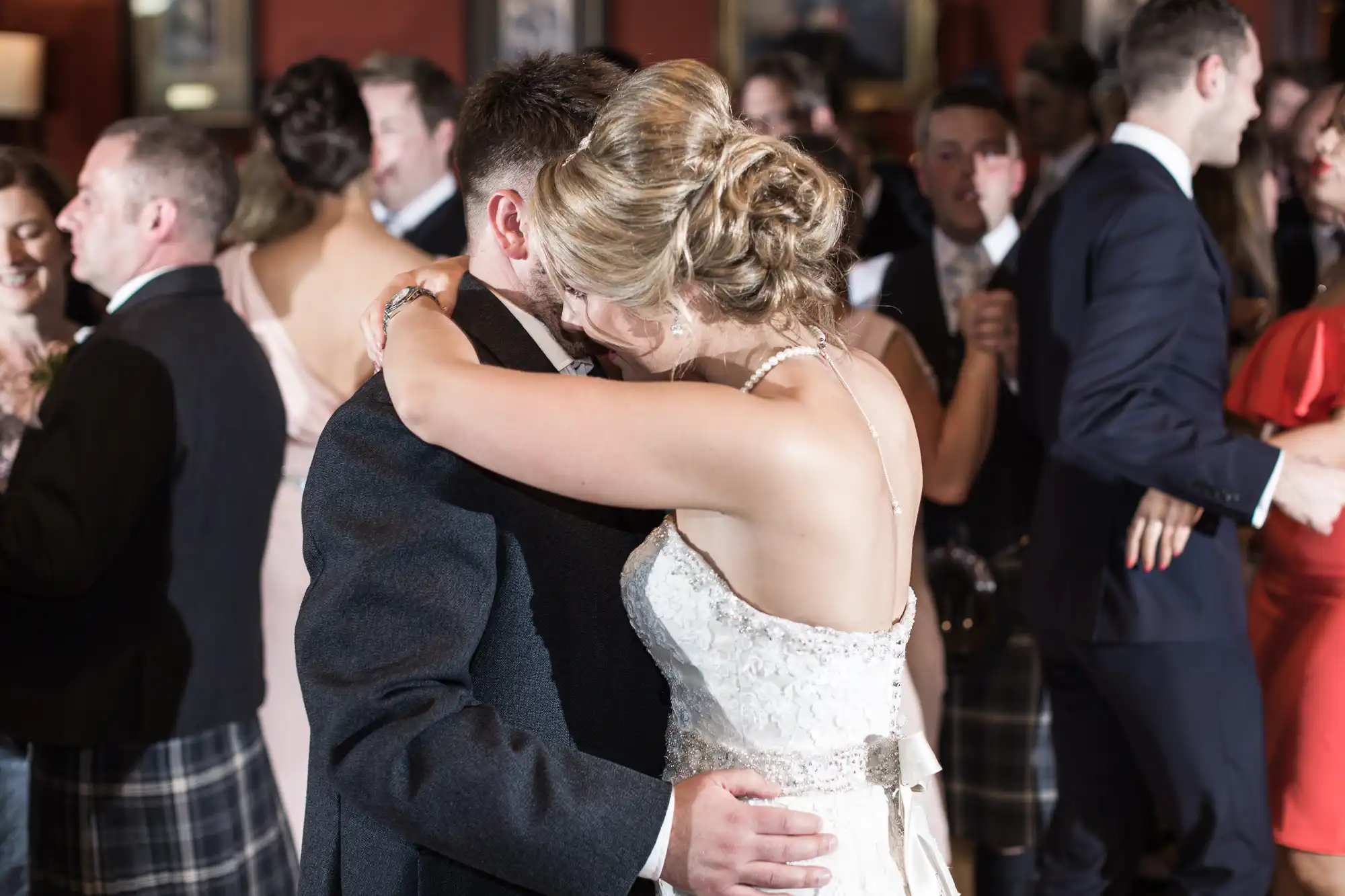 A couple embraces and dances in formal attire at a crowded indoor event. Other attendees are engaged in conversation or dancing in the background.