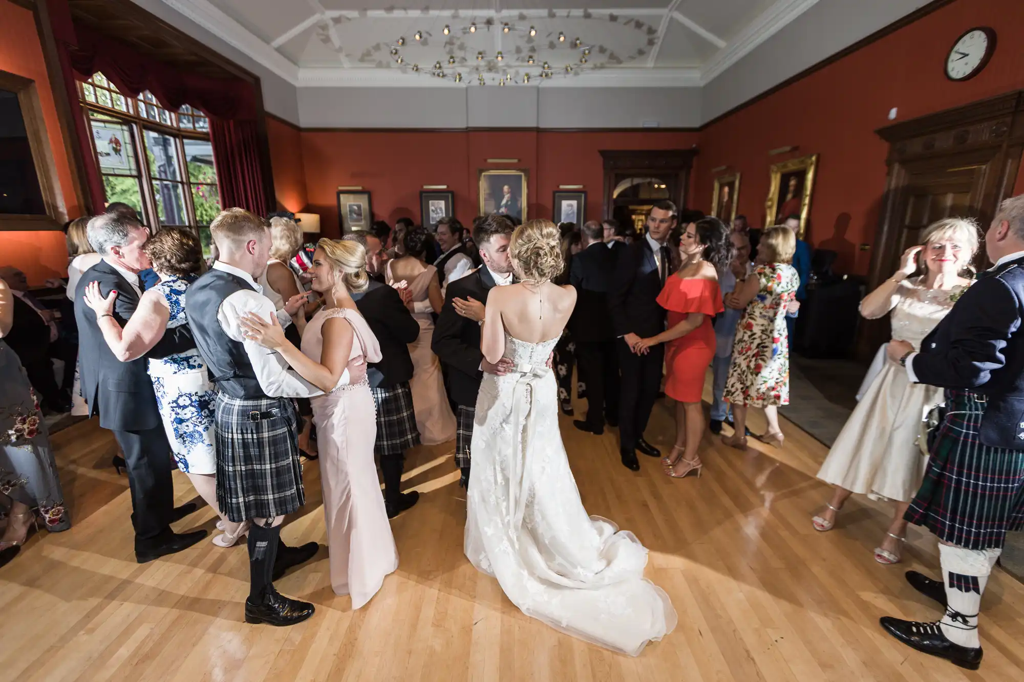 A group of people are dancing in a room with red walls and wooden accents. A bride in a white gown is visible in the center. Several men wear kilts, and some guests are dressed in formal attire.