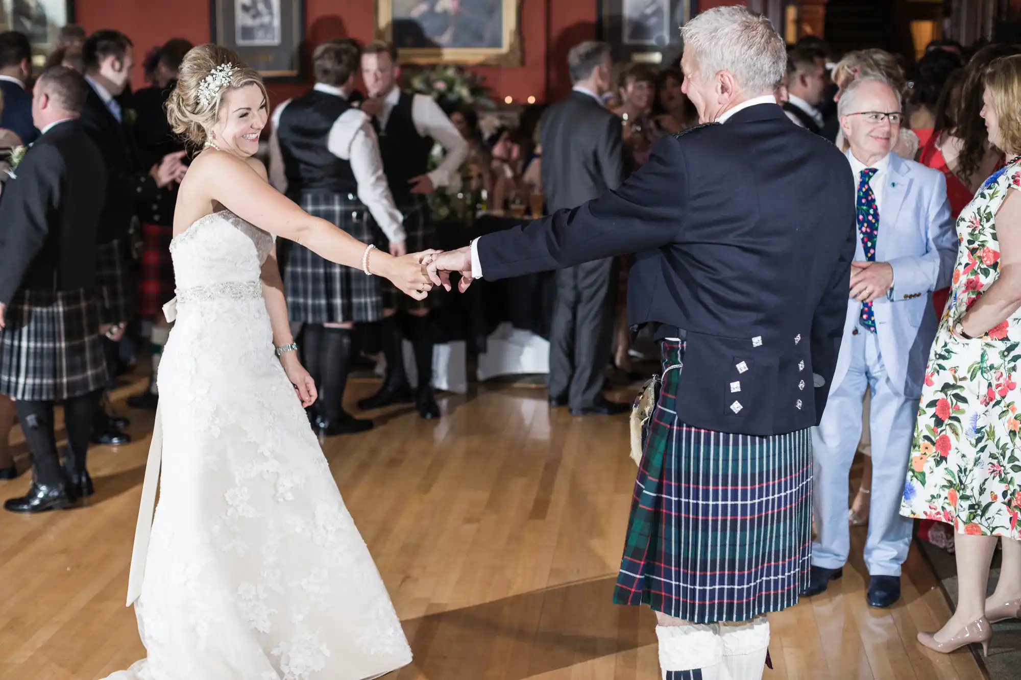 A bride and an older man in traditional Scottish attire dance while guests watch.