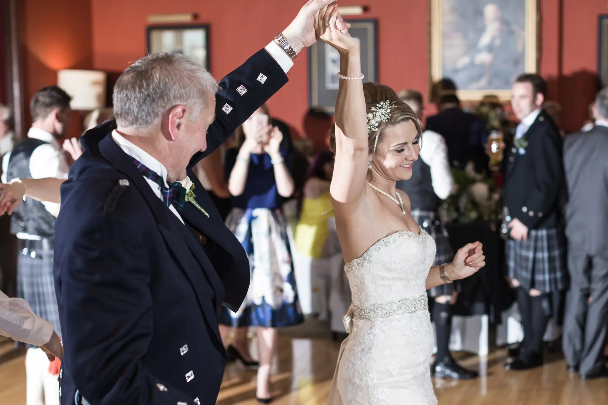 A bride and a man in formal attire dance together at a wedding reception, while guests observe and chat in the background.