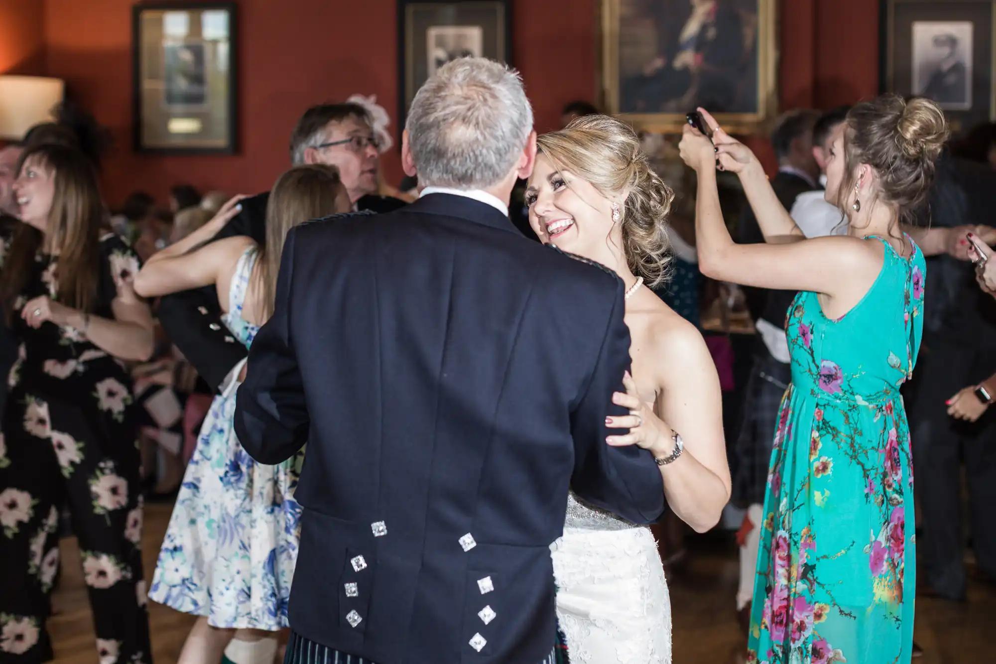 A couple dances closely at a lively event, surrounded by other people dancing. The room has warm lighting and framed pictures on the walls.