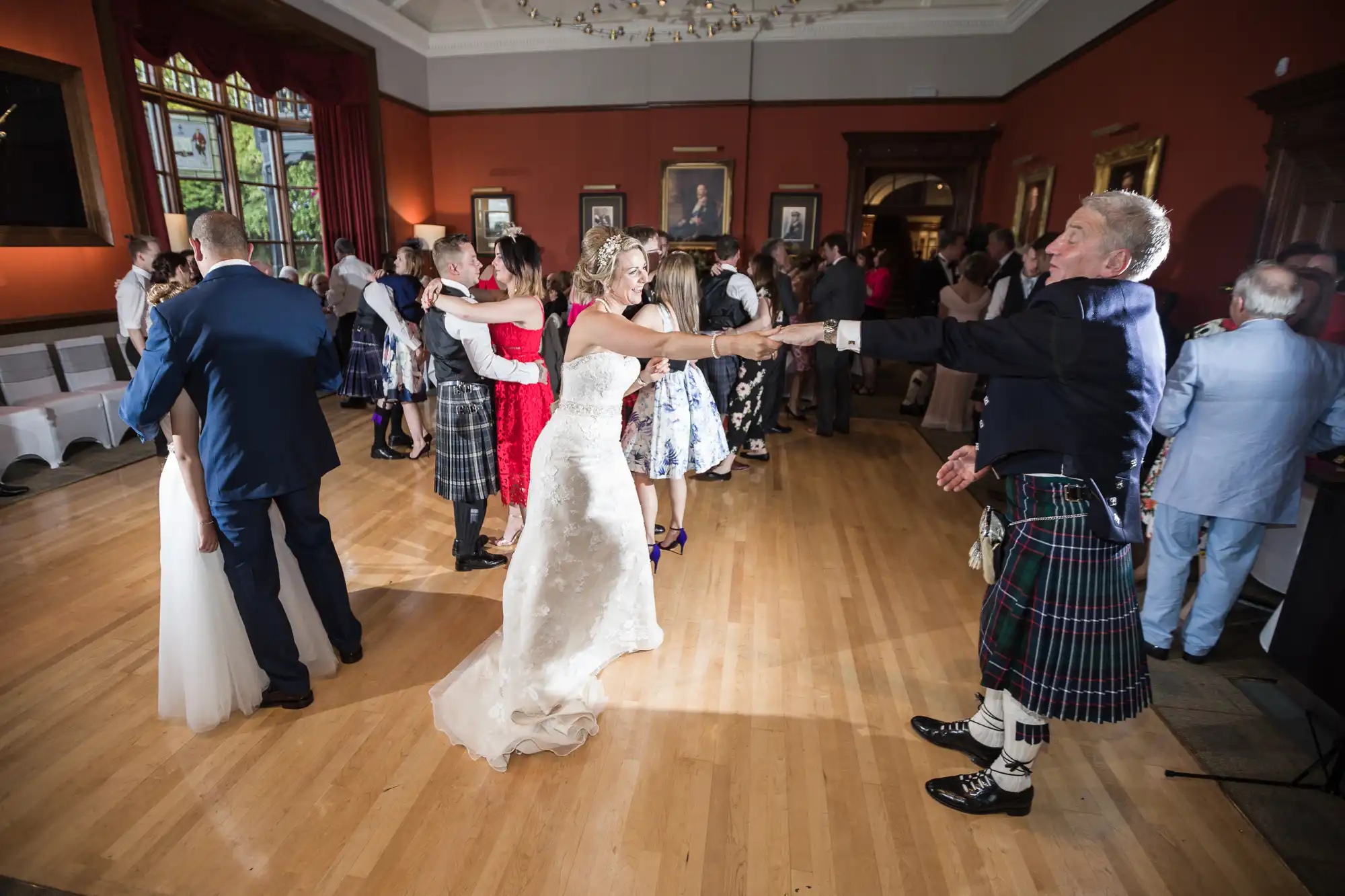 A bride and groom in traditional Scottish attire dance in a brightly lit room with wooden floors, surrounded by other couples also dancing.