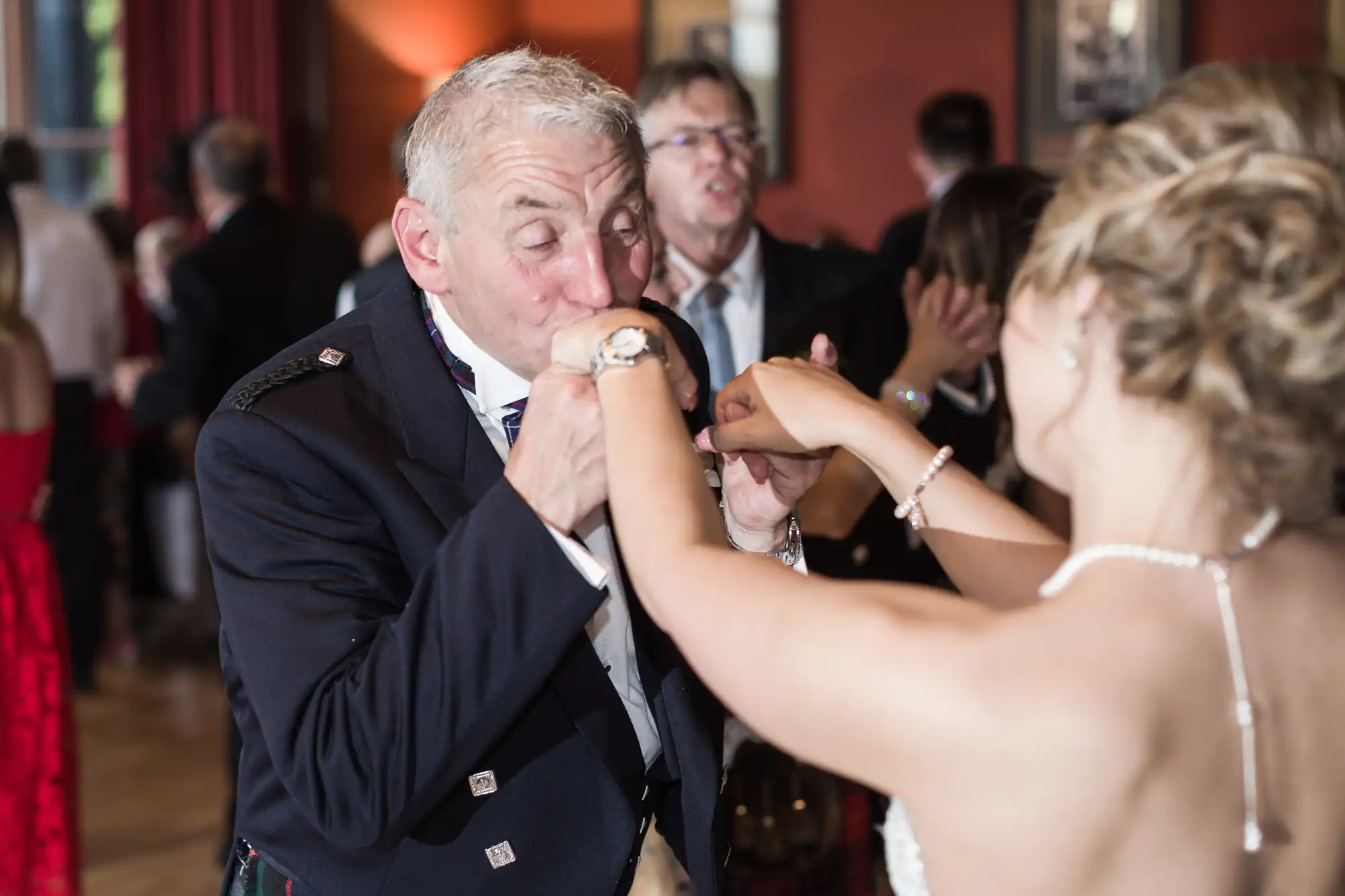 An elderly man wearing a suit kisses the hand of a woman in a strapless dress at a formal event, with other attendees and elegant decor visible in the background.