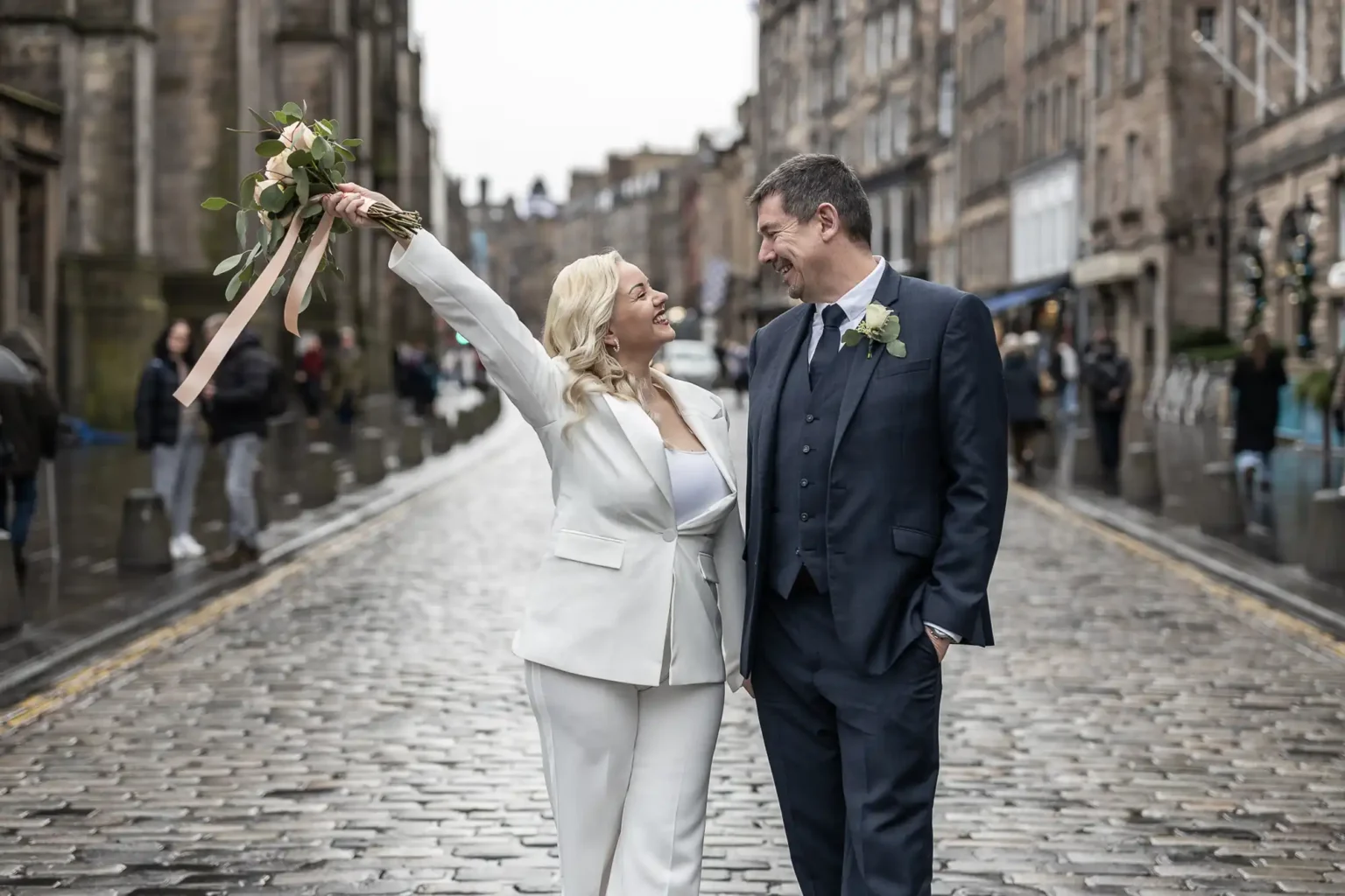 A woman in a white suit holds a bouquet and smiles at a man in a dark suit as they stand together on a wet cobblestone street, surrounded by historic buildings.