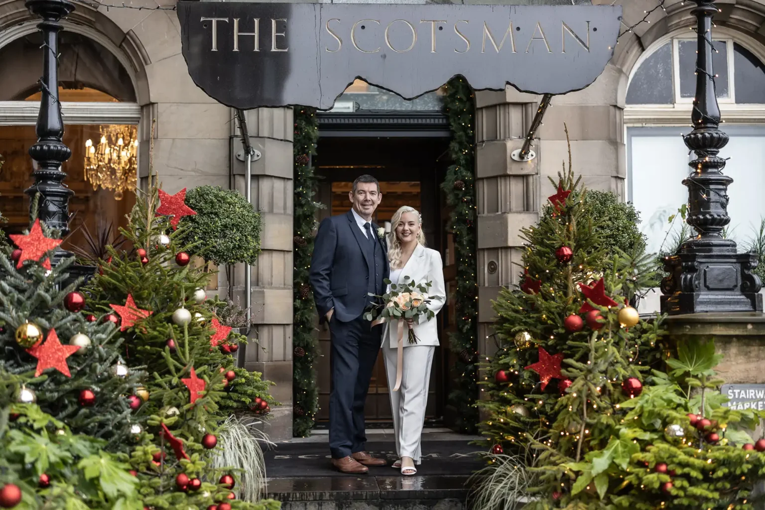 A couple dressed formally stands outside The Scotsman building, surrounded by decorated Christmas trees and festive greenery.