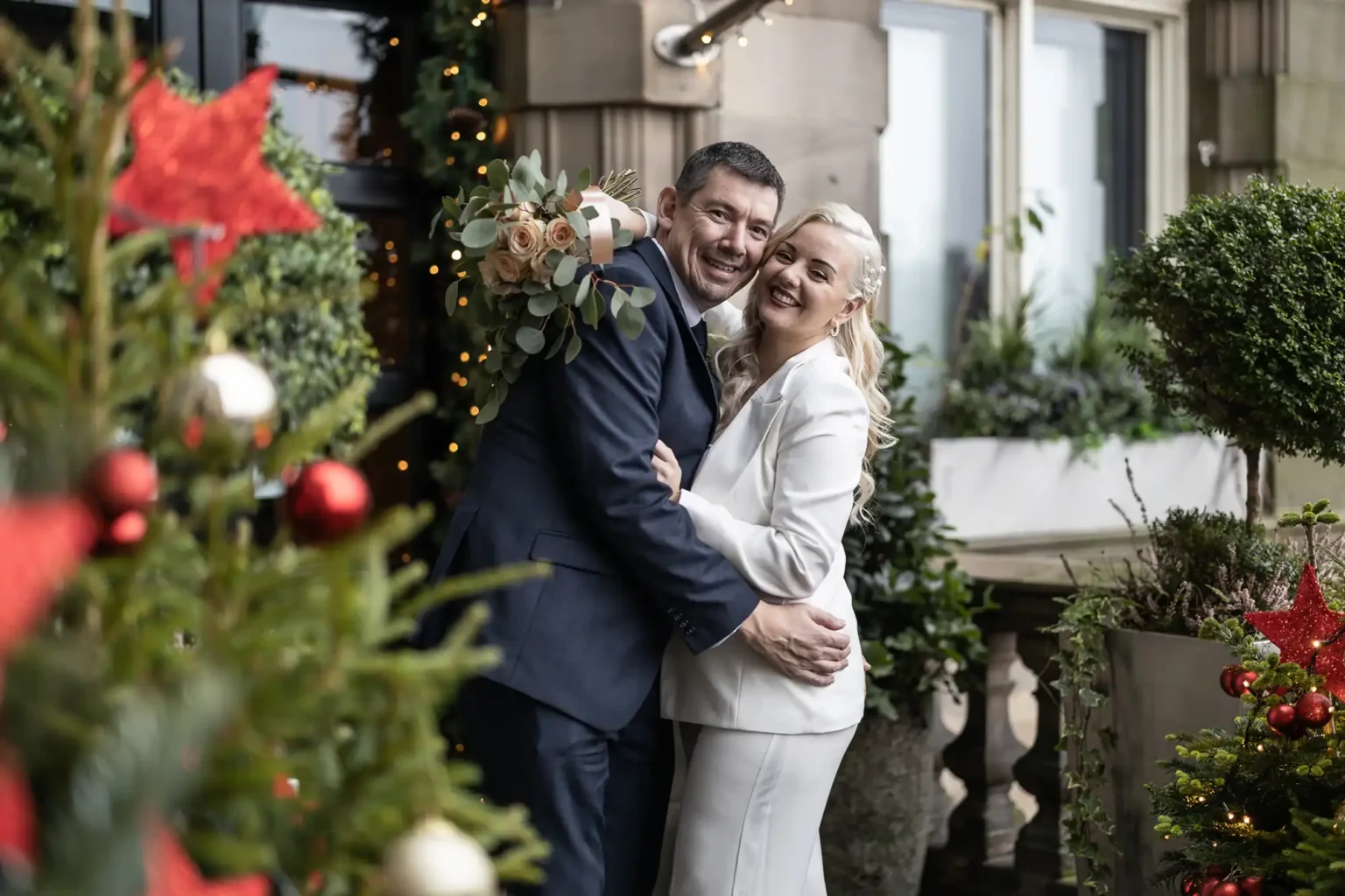 A couple dressed formally, smiling and embracing outdoors, surrounded by festive Christmas decorations and greenery.