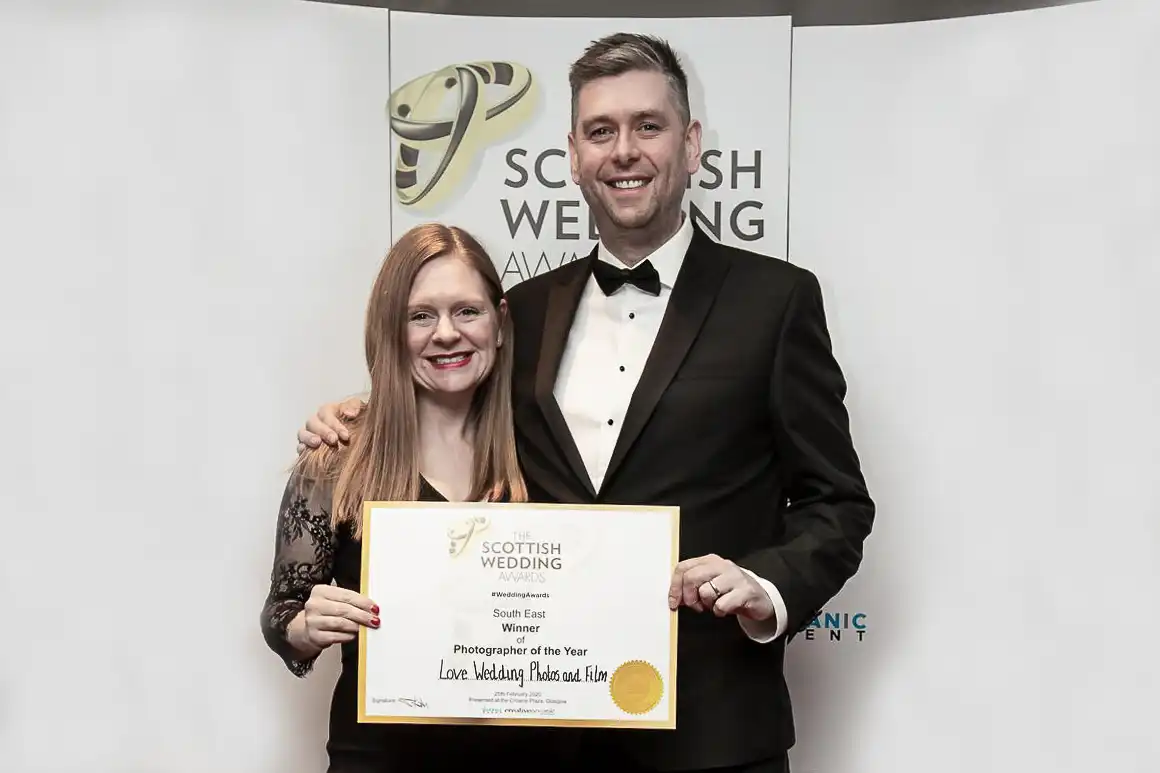 Claire and Jonathon dressed formally stand on a red carpet holding an award certificate in front of a backdrop with multiple logos at the Scottish Wedding Awards awards ceremony.