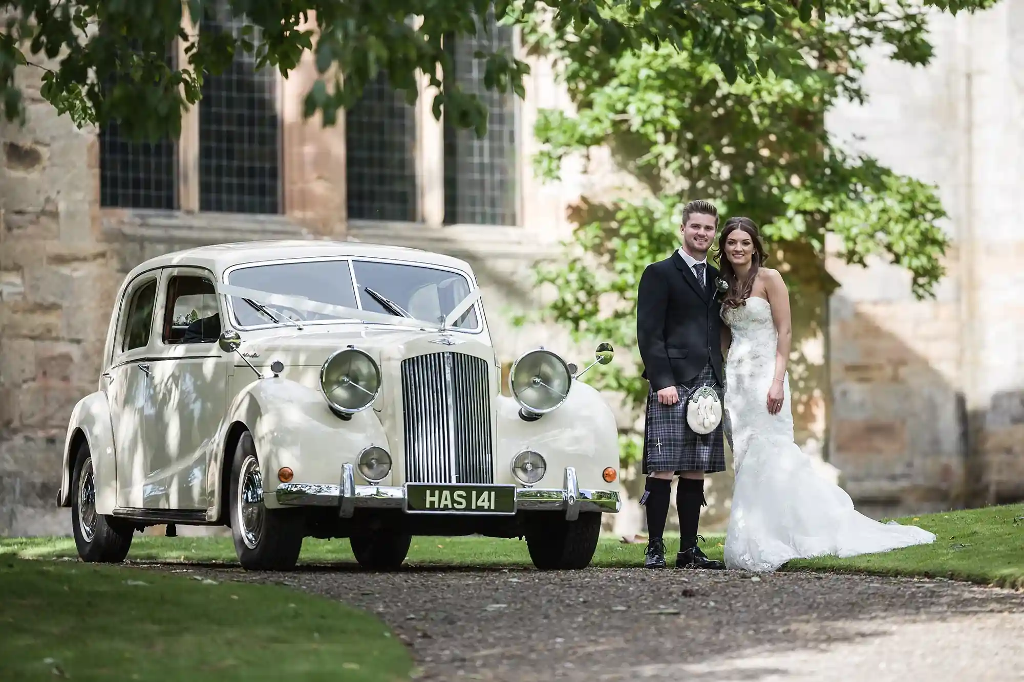 A newlywed couple stands beside a classic white vintage car, with the groom in a kilt and the bride in a long white gown, in front of a historical stone building.
