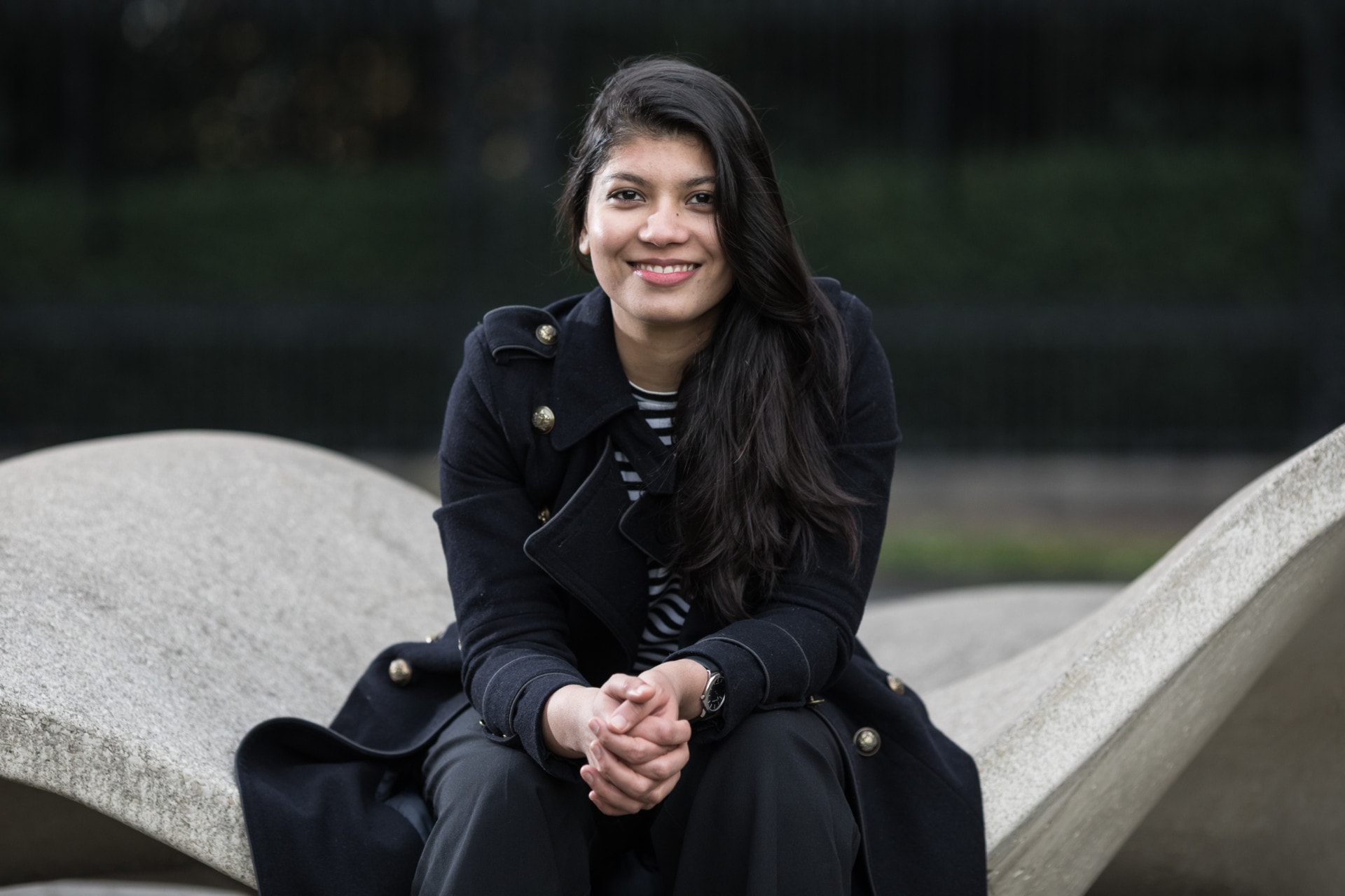 A young woman in a black coat sitting on a concrete structure, smiling at the camera.