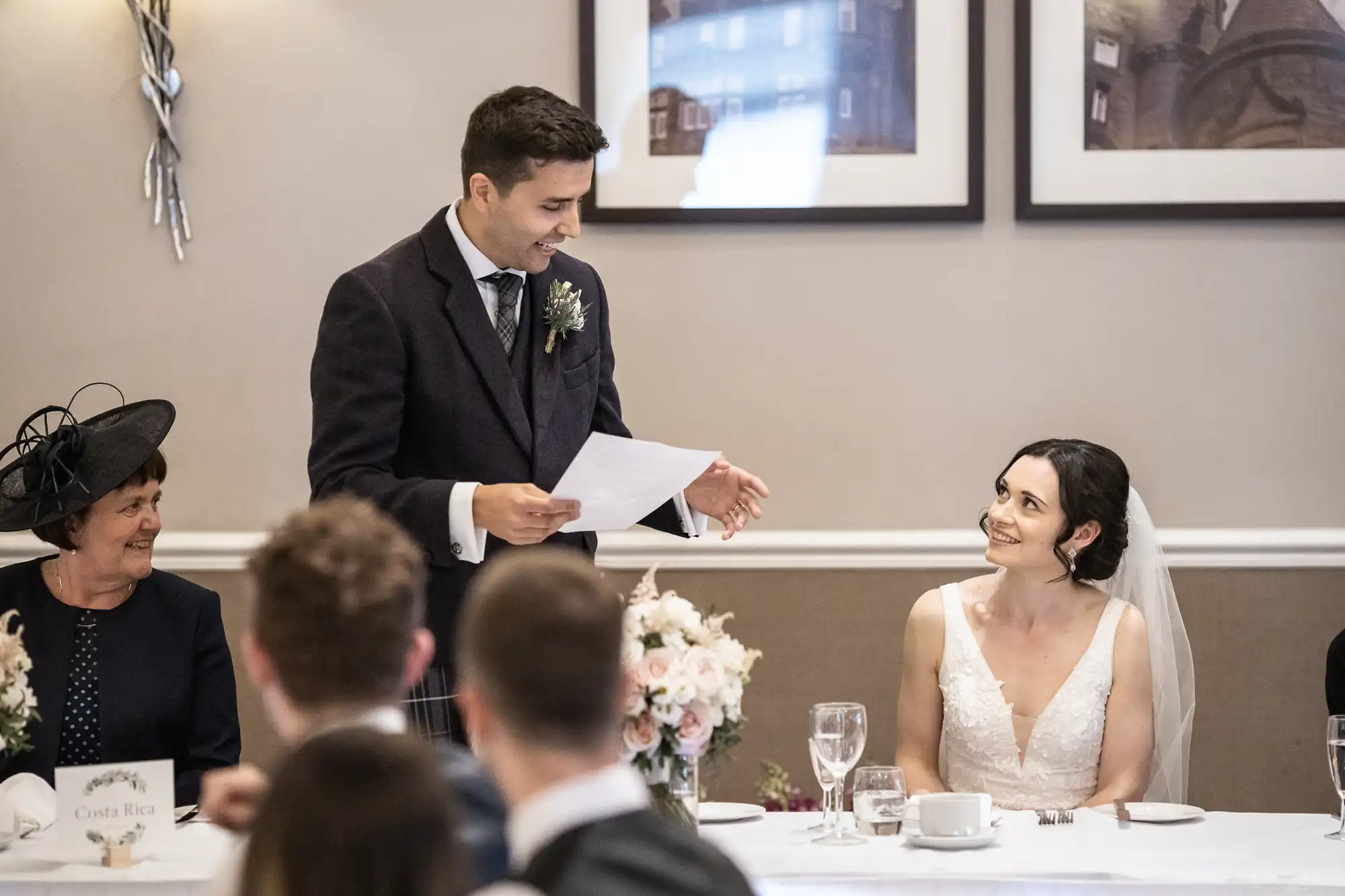 A person stands giving a speech at a wedding reception, holding a paper, while another person in bridal attire sits smiling. Guests are seated nearby.
