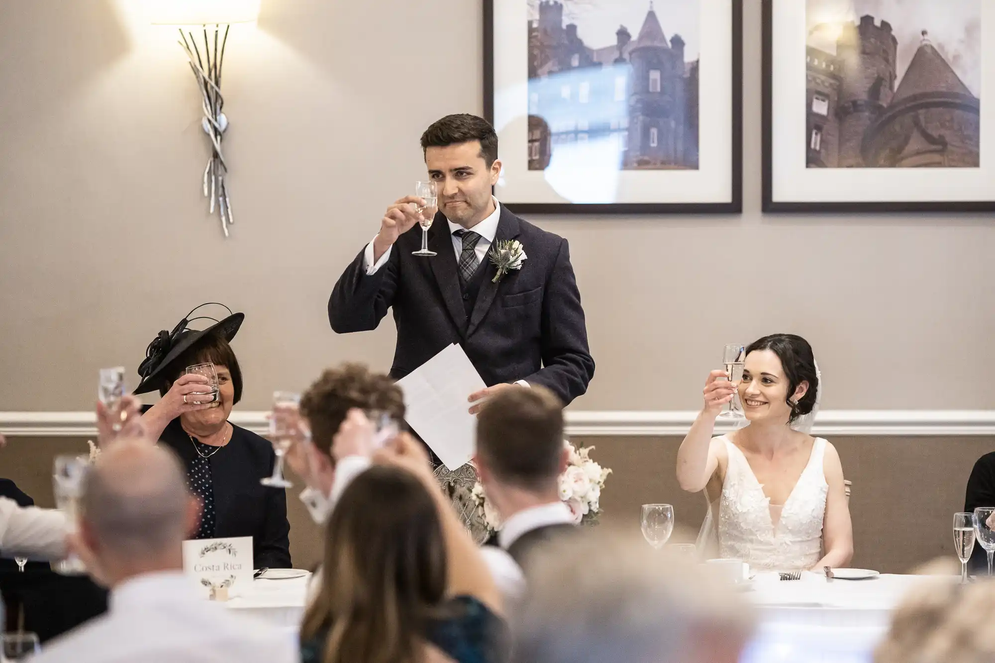 A man in a suit delivers a speech holding a champagne glass, while guests, including a woman in a white dress, raise their glasses in a toast during a formal event.