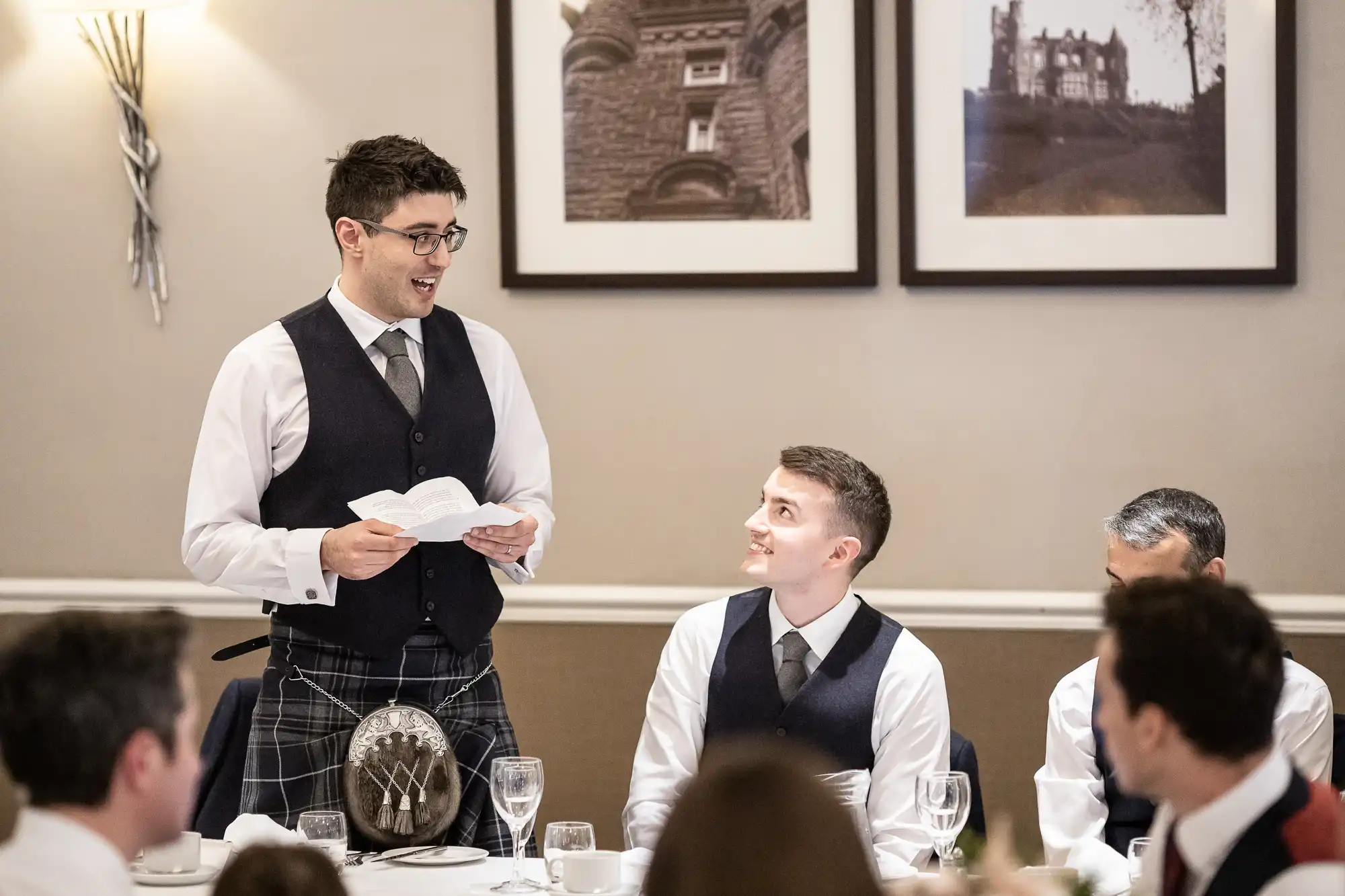 A man in a vest gives a speech at a formal event while others seated at the table listen. Two framed photos are on the wall behind them.
